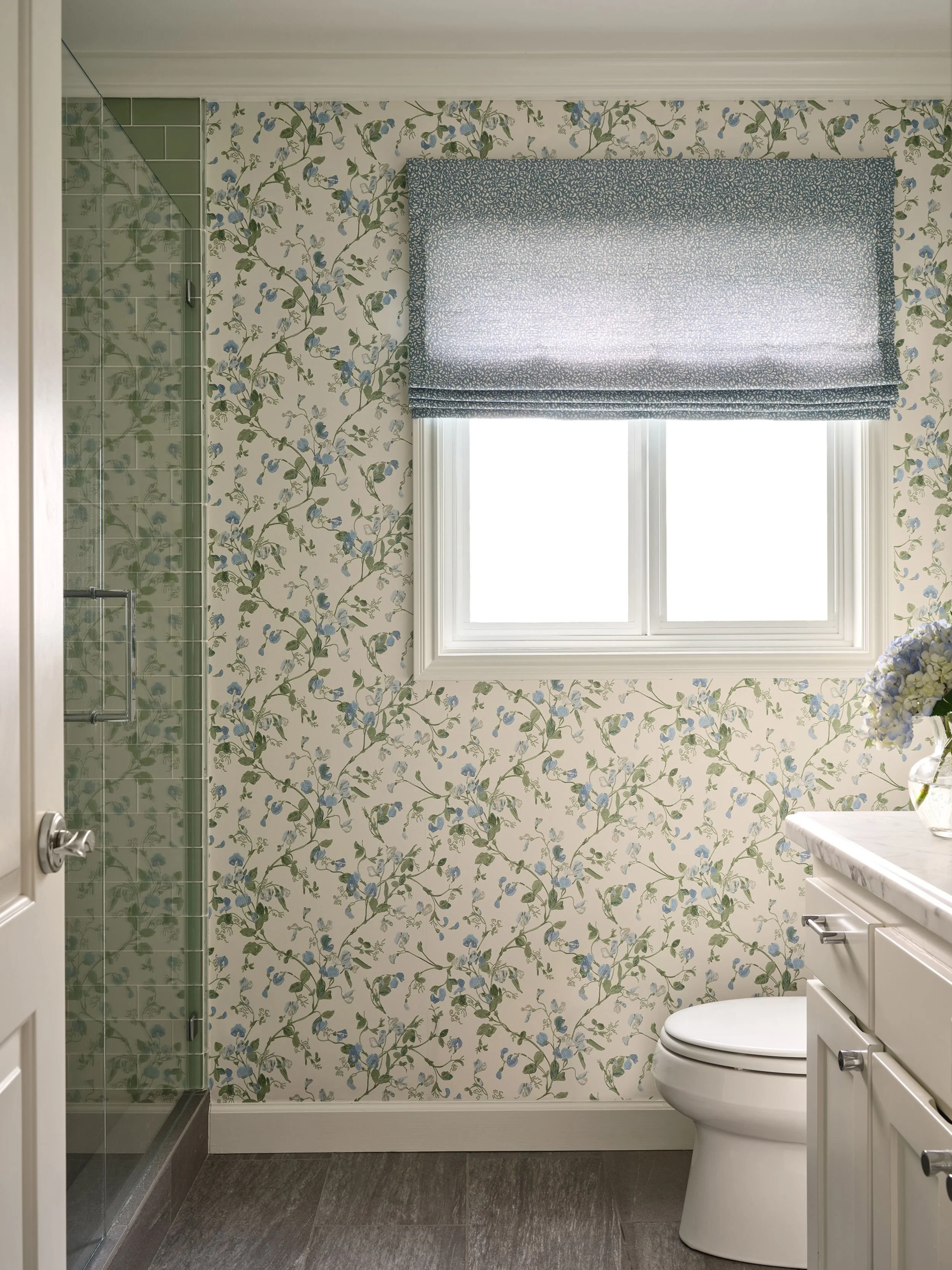Bathroom with floral wallpaper, a window with a blue patterned Roman shade, a glass shower enclosure, a white toilet, and a white vanity with a marble countertop and a vase of blue flowers.