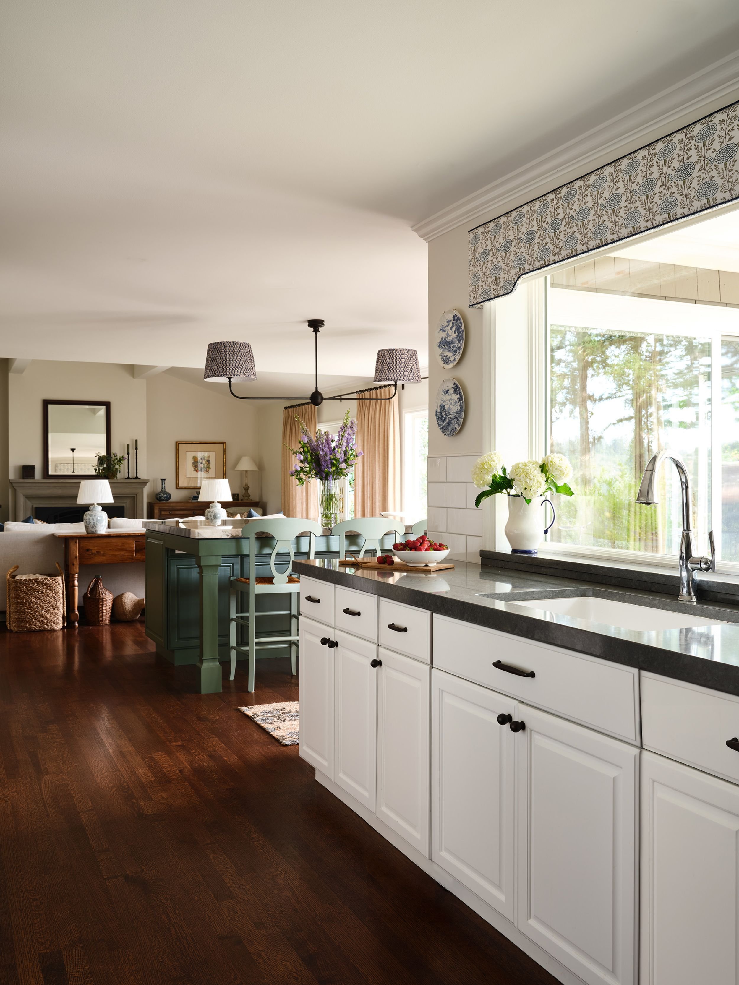 Kitchen and dining area with white cabinets, dark countertops, large window, flowers, cherry tomatoes, and a view of trees outside.