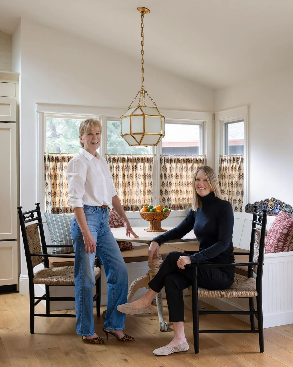 Two women in a bright kitchen with large windows and patterned curtains, one standing and one seated at a wooden dining table with a fruit bowl, smiling at the camera.
