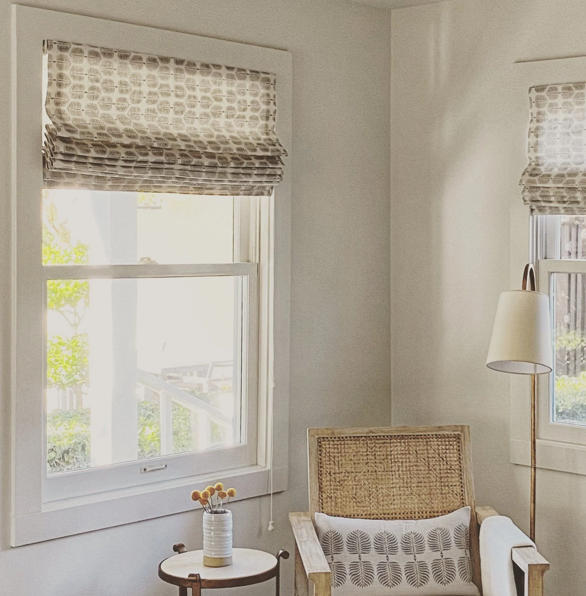 A cozy corner of a room with two windows dressed in patterned Roman shades, a wooden chair with a decorative cushion, a small round side table with a vase holding yellow flowers, and a tall floor lamp with a white shade.