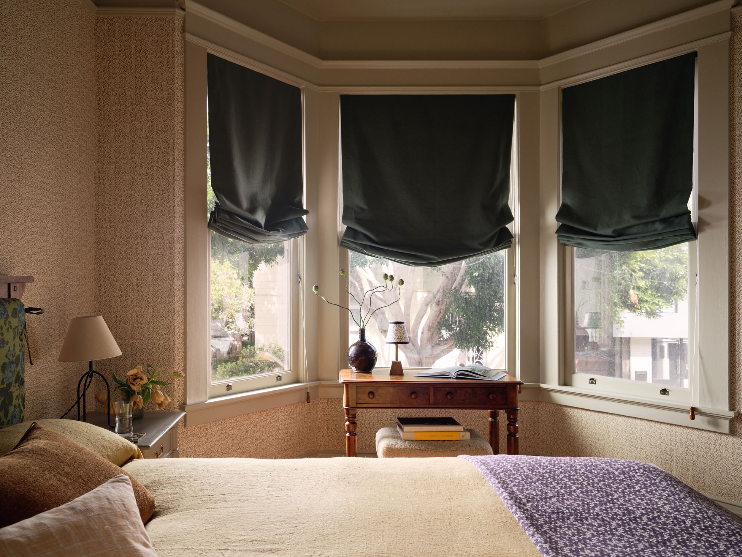 A cozy bedroom with a bed in the foreground, a nightstand with a lamp and flowers to the left, and a wooden table with a vase, lamp, and an open book in front of three bay windows with dark roman shades. Outside the windows are trees and neighboring 