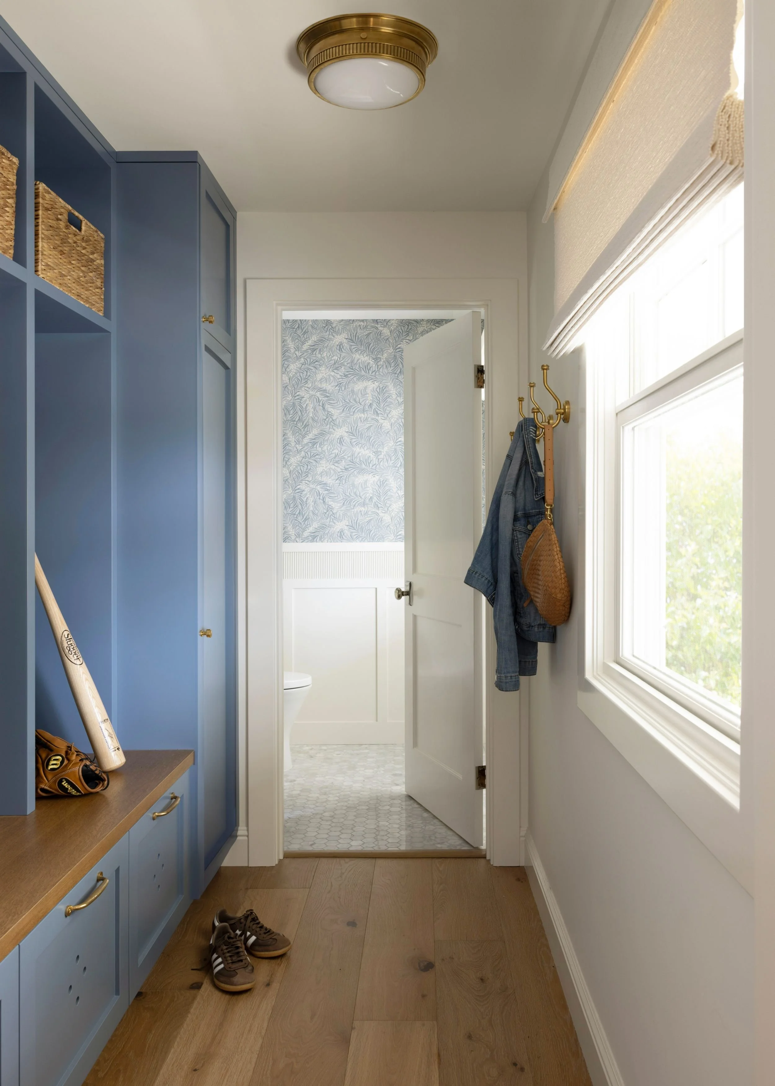 Entryway with blue built-in shelves, baseball bat, baseball glove, brown shoes on wooden floor, coat hooks with denim jacket and brown bag, window with blinds, open door leading to a bathroom with blue leaf-patterned wallpaper.