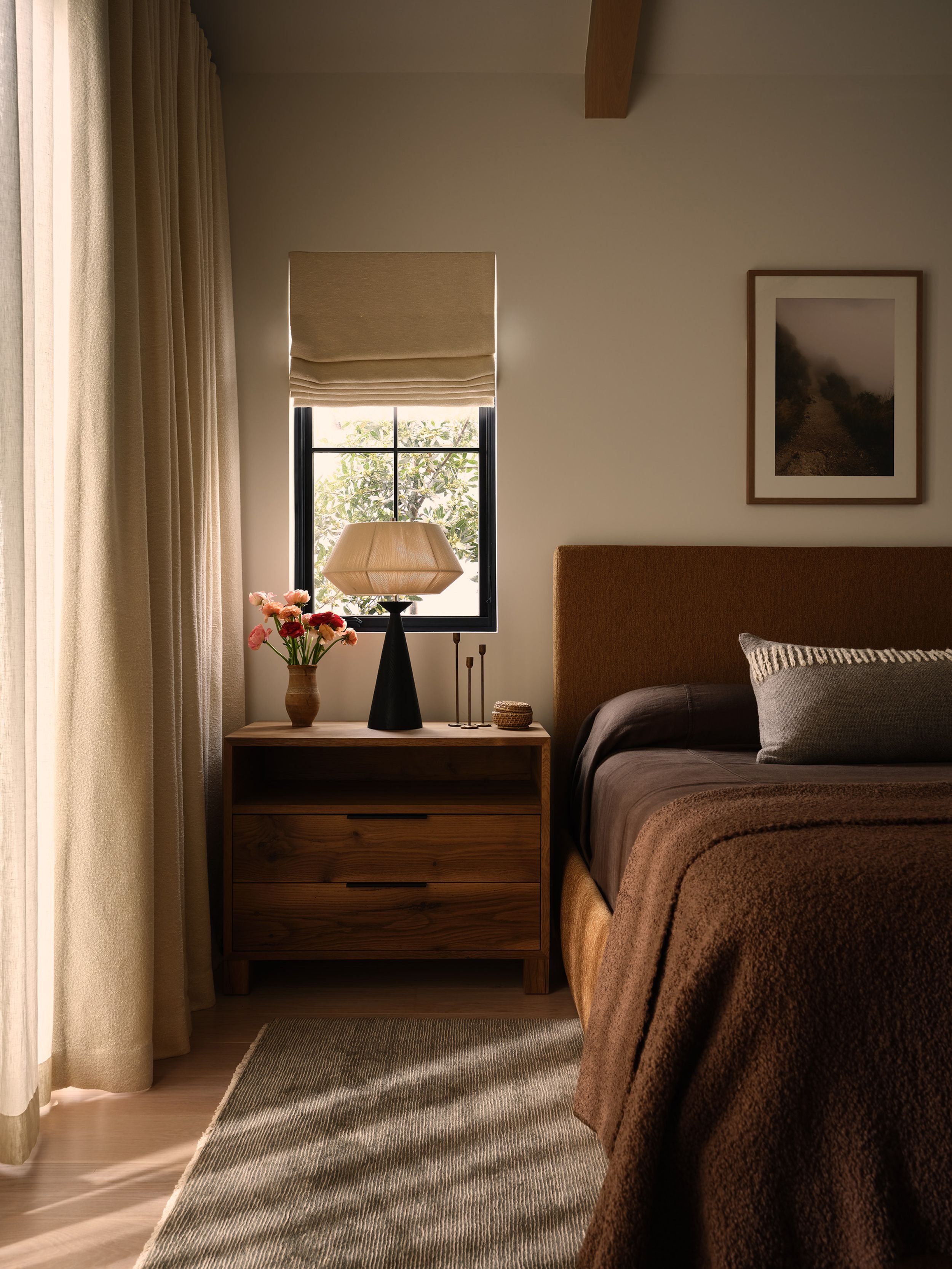 A cozy bedroom with a wooden bedside table holding a black and beige table lamp and a vase of pink flowers, next to a window with beige curtains, and a framed landscape photograph on the wall above the bed's headboard.