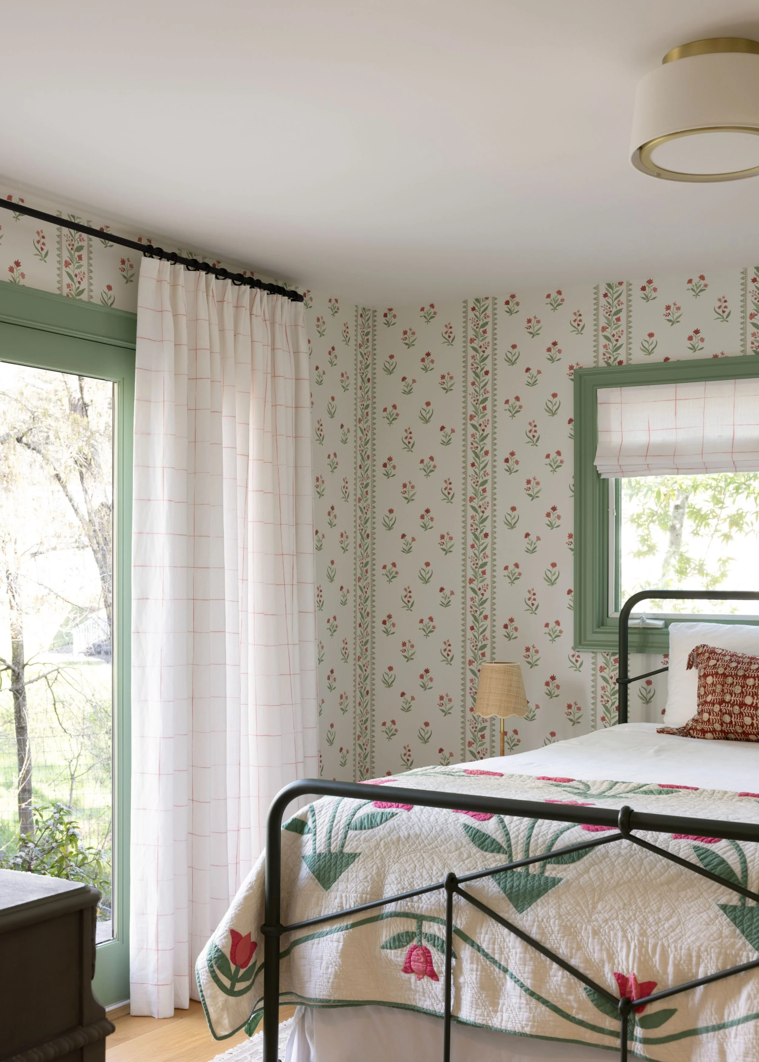 A bedroom with floral wallpaper, a bed with a quilted coverlet featuring pink flowers, a window with green trim, white curtains, and a black metal bed frame.