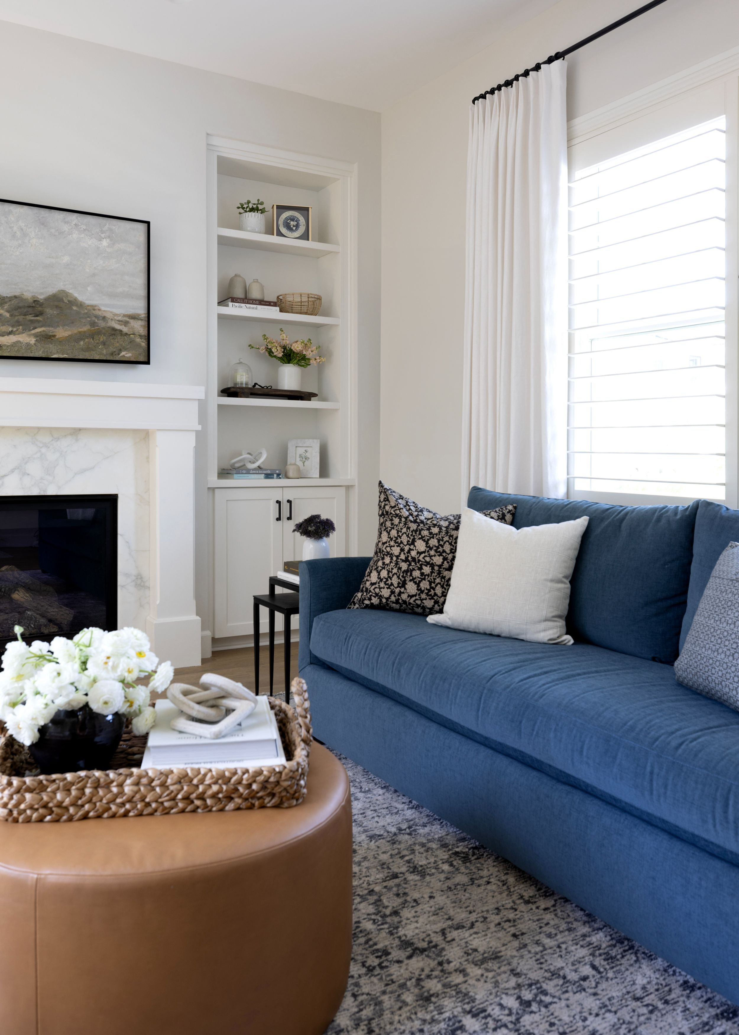 Living room with a blue sofa, decorative pillows, a coffee table with flowers, a fireplace, and white curtains.