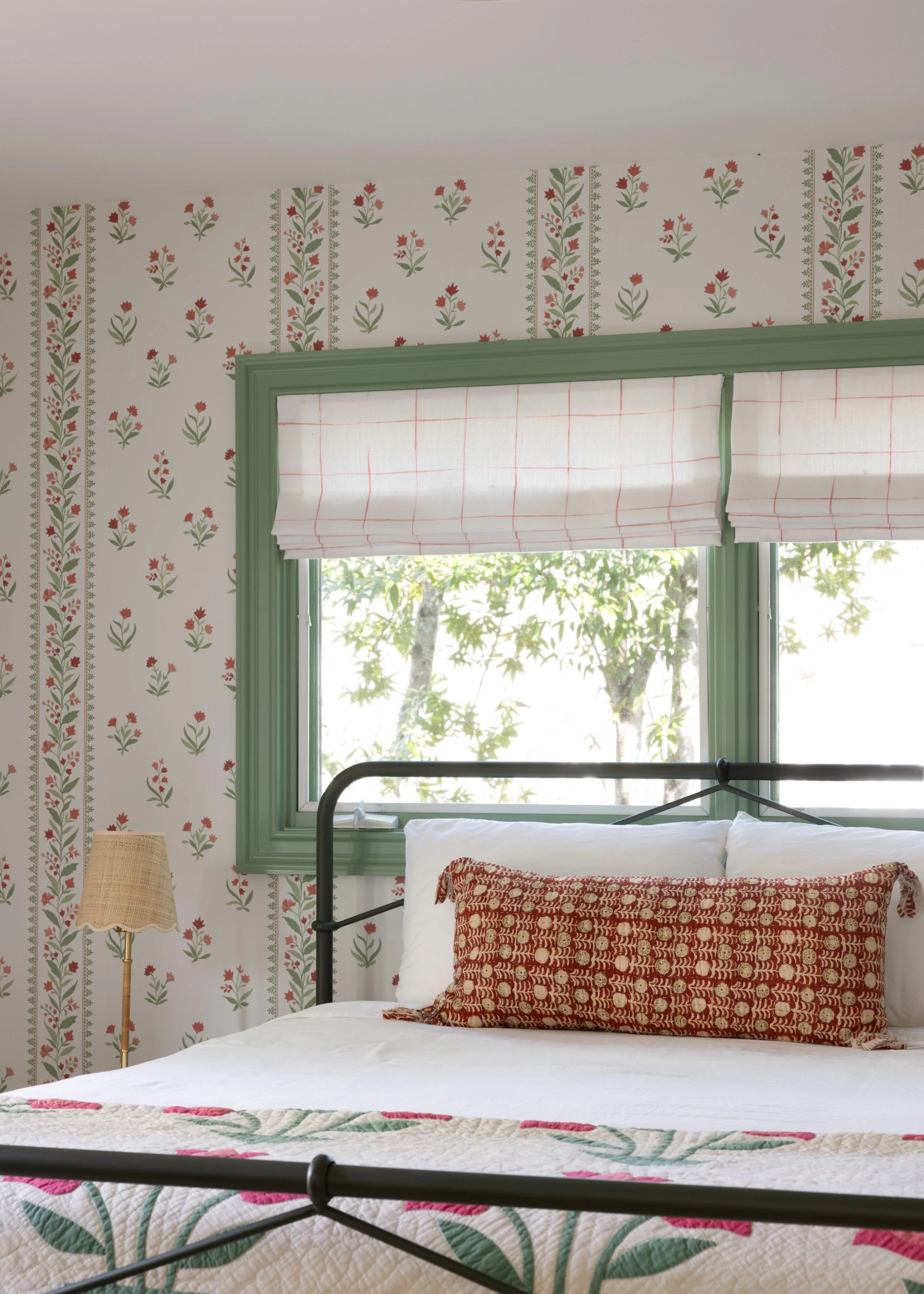 A bedroom with a bed, patterned quilt, decorative pillow, and a window with greenery outside. The walls have floral wallpaper, and there is a white Roman shade on the window.
