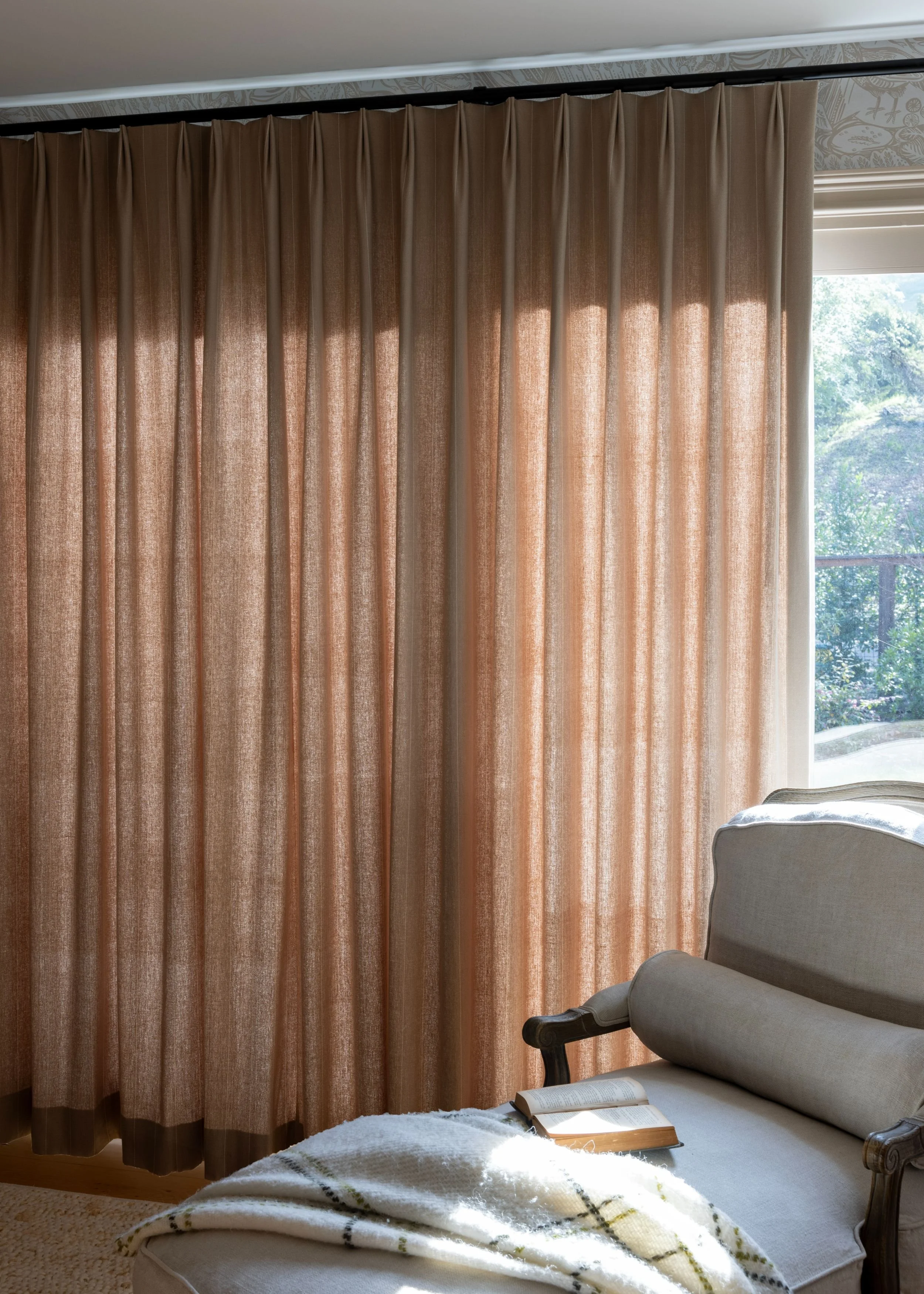 Sunlight filters through light brown curtains in a living room, revealing a glimpse of trees outside a window. A beige upholstered armchair with a rolled pillow on top sits next to the window, with an open book and a plaid blanket on the armrest.