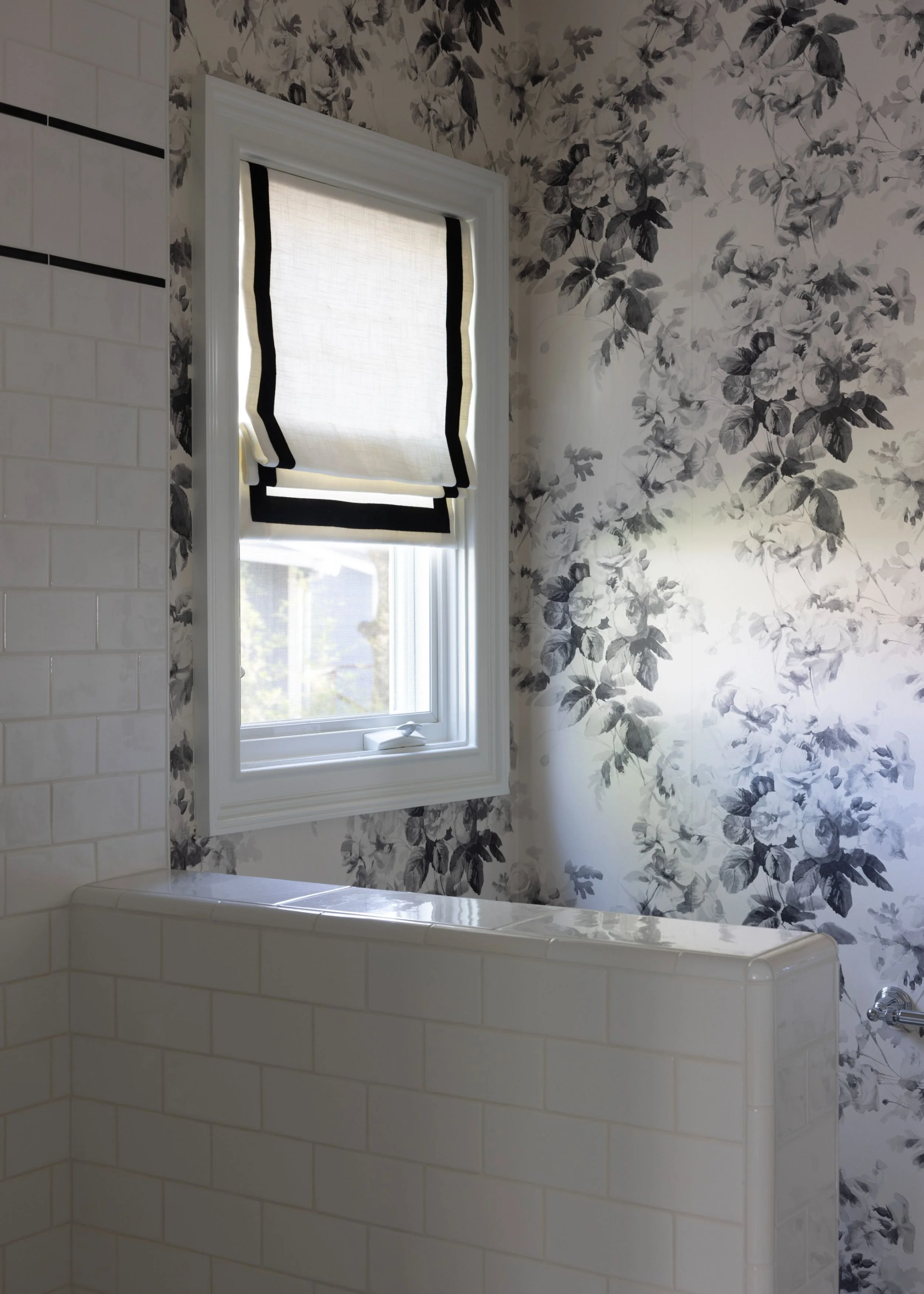 A bathroom with a window covered by a white Roman shade with black trim. The wall features black and white floral wallpaper, and there are white subway tiles on the nearby wall.
