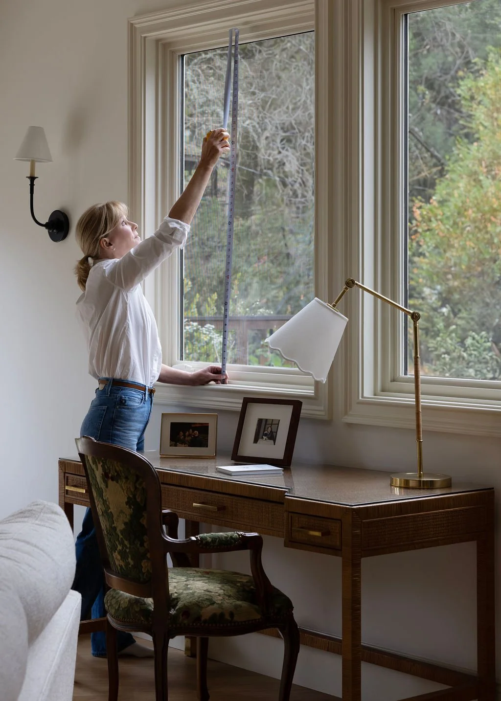 A woman measuring the height of a window with a measuring tape inside a room with a desk, a lamp, and framed photos, with trees visible outside.