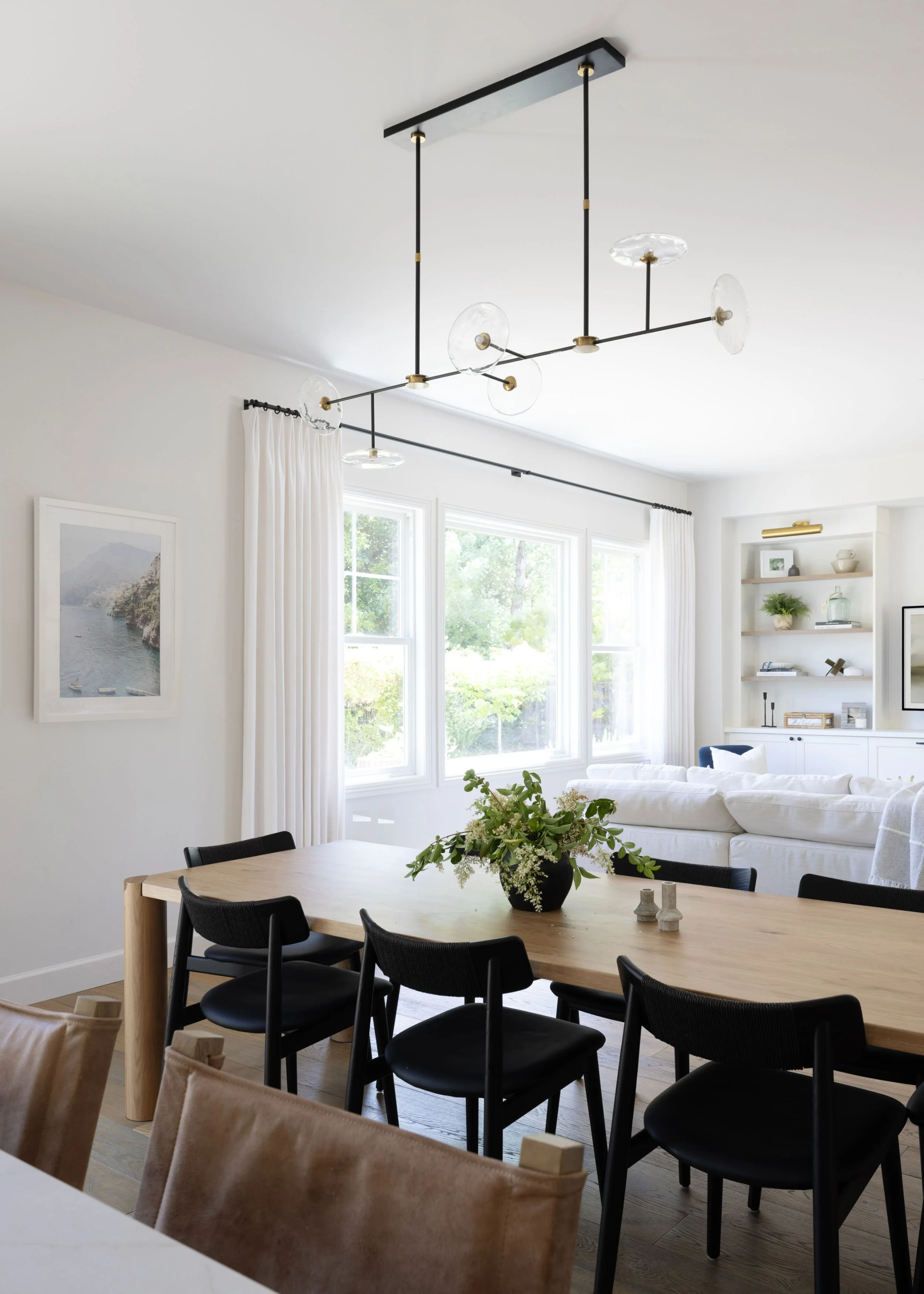 A modern dining and living room with natural light, black dining chairs, a wooden table, a black vase with flowers, white curtains, wall art, built-in white shelves with decor, and a black and gold ceiling light fixture.