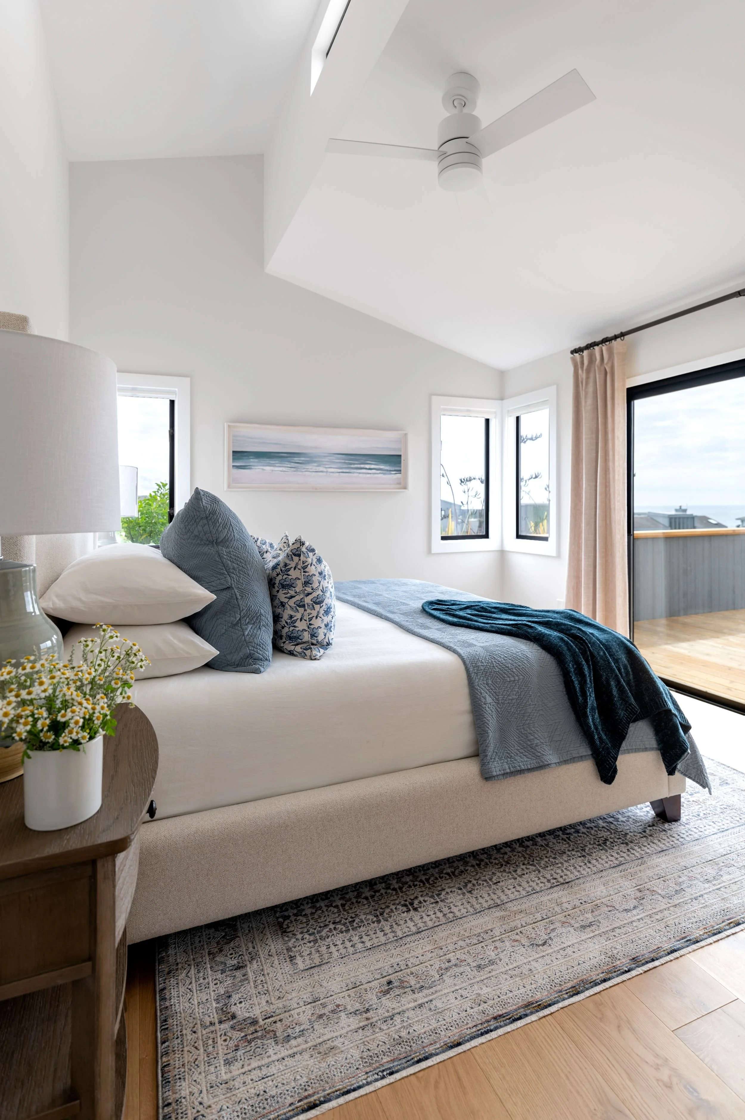 A cozy bedroom with a bed that has white sheets and blue and floral pillows. There is a wooden nightstand with a white lamp and a potted plant. The room has white walls, large windows, and a sliding glass door leading to a balcony, with a ceiling fan