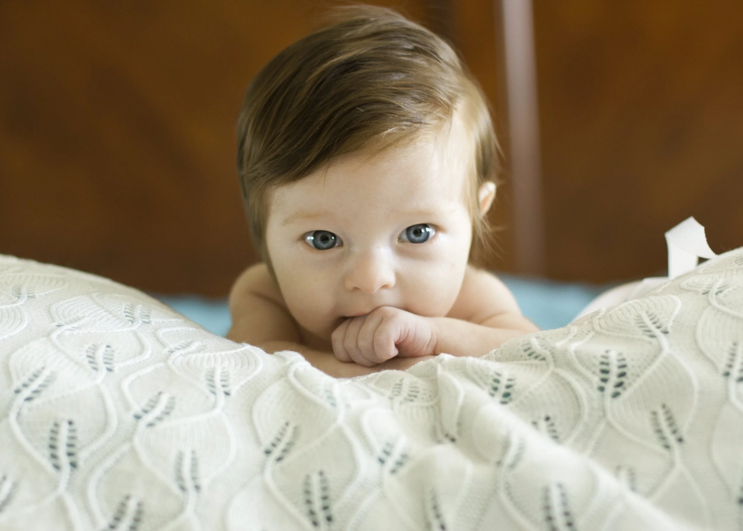 A young child with blue eyes and brown hair, lying on a bed with a white textured blanket, looking directly at the camera.