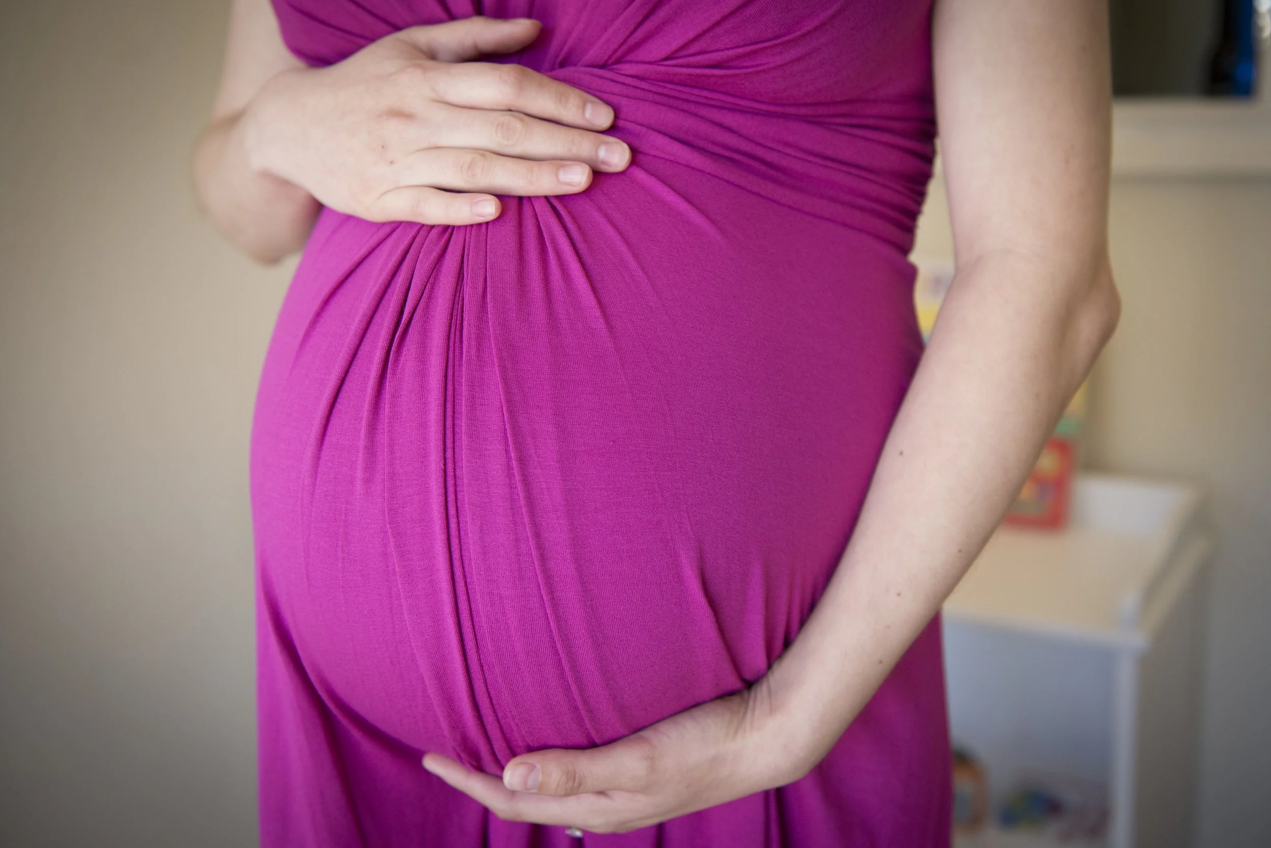 A woman in a pink dress holds her pregnant belly with one hand resting on top and the other underneath.