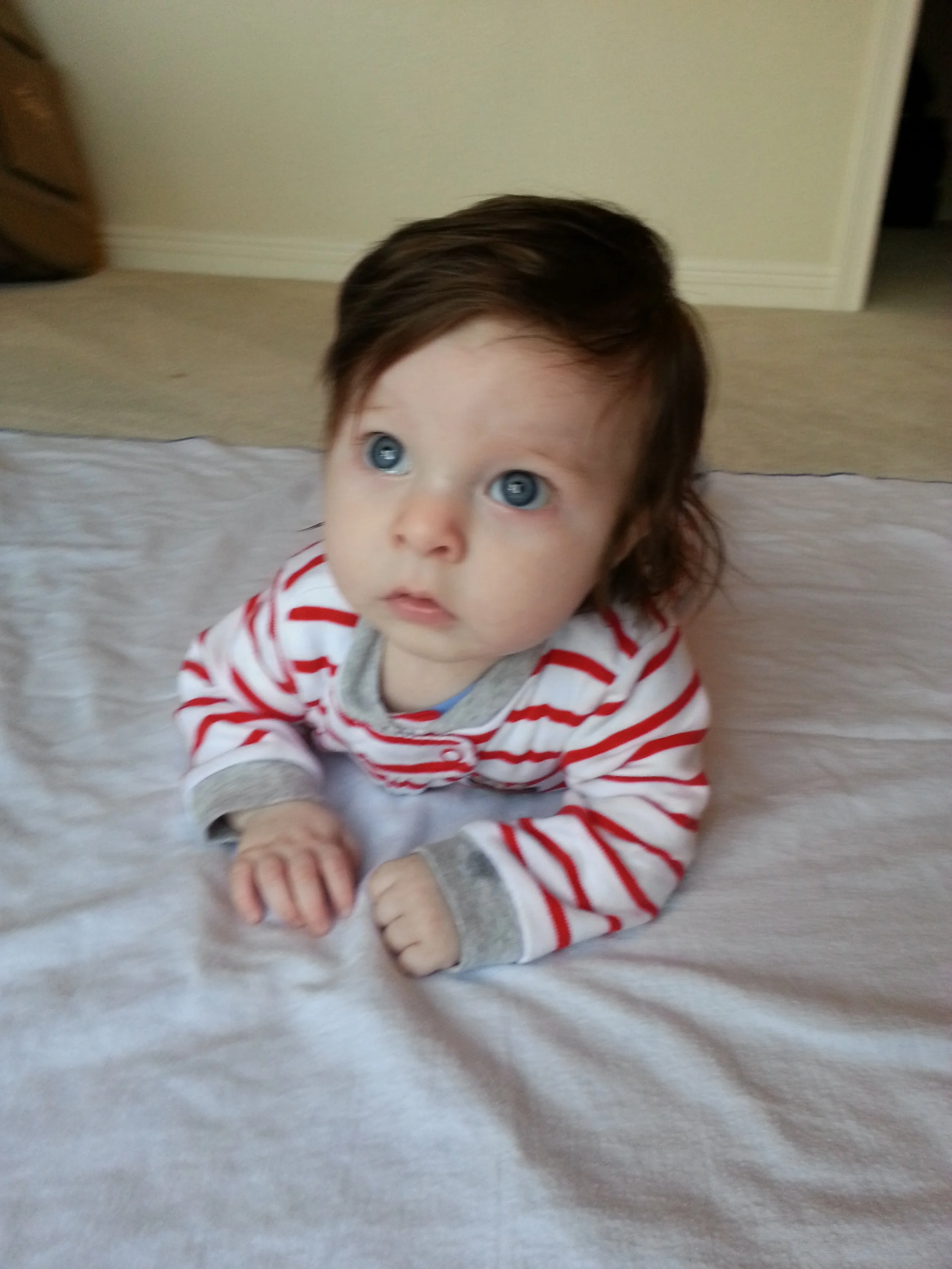 A baby with blue eyes and dark hair lying on a soft surface, wearing a white and red striped shirt.