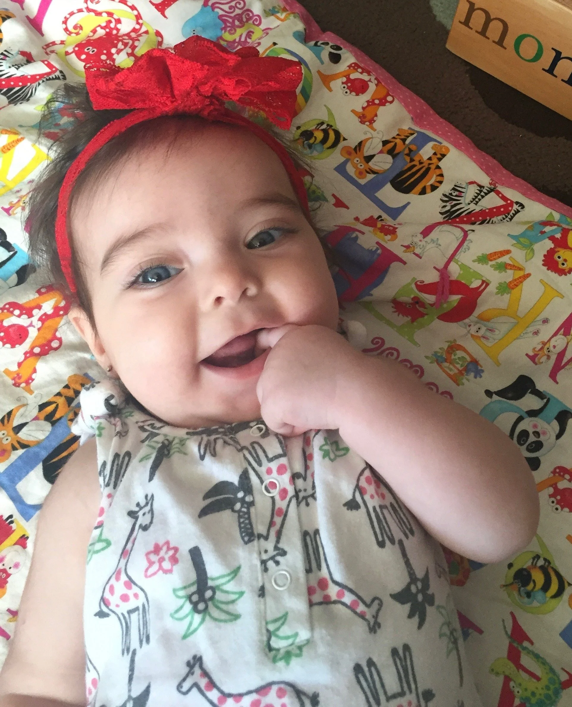 A smiling baby girl with blue eyes, wearing a sleeveless shirt with zebra and giraffe prints, lying on a colorful blanket with animal illustrations, and wearing a red bow headband.