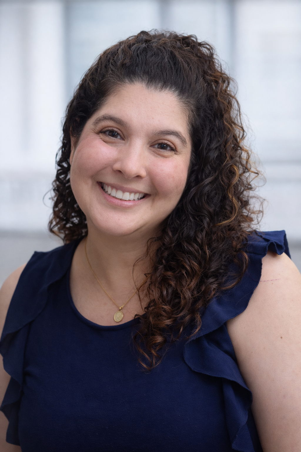 A woman with curly dark hair, smiling, wearing a navy blue sleeveless top with ruffled sleeves and a gold necklace with a pendant.