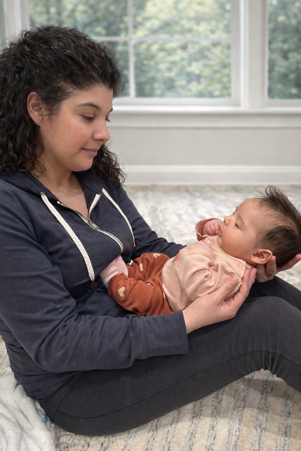 A woman sitting on the floor holding a baby in her lap, looking at the baby with affection, in a room with large windows and natural light.