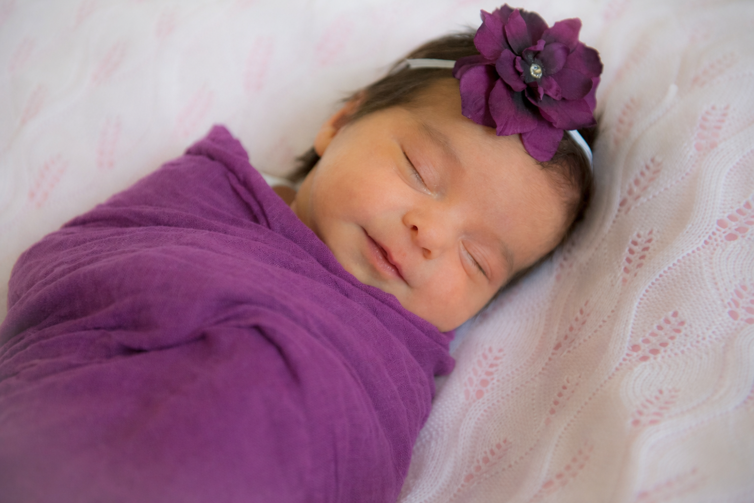 A young girl sleeping peacefully, wrapped in a purple blanket, with a purple flower headband on her head, lying on a soft, patterned bed.