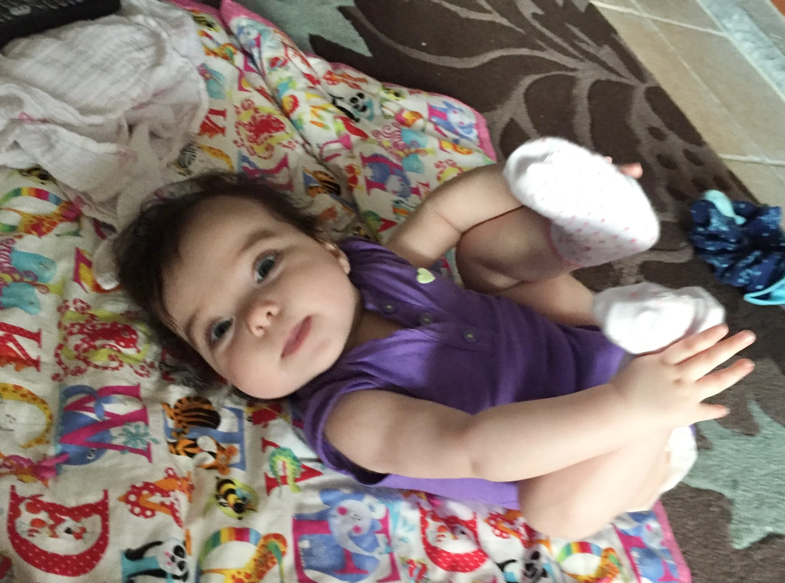 A young child lying on a bed with a colorful animal-themed bedsheet, wearing a purple shirt and white socks with pink polka dots, looking up at the camera.