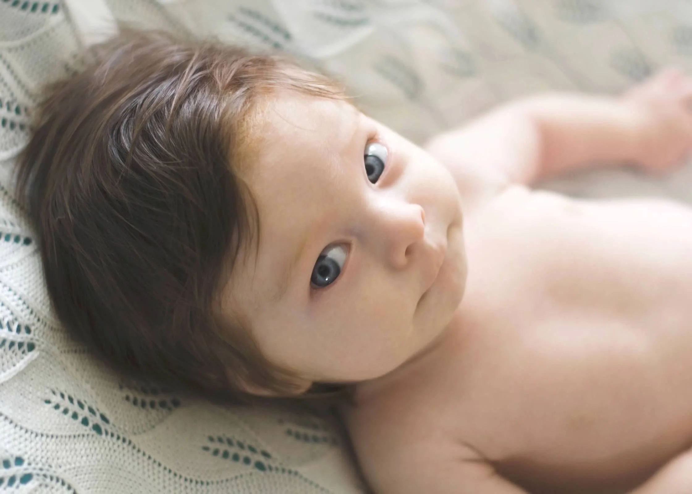 Close-up of a baby lying on a woven surface, looking towards the camera with wide blue eyes and brown hair.