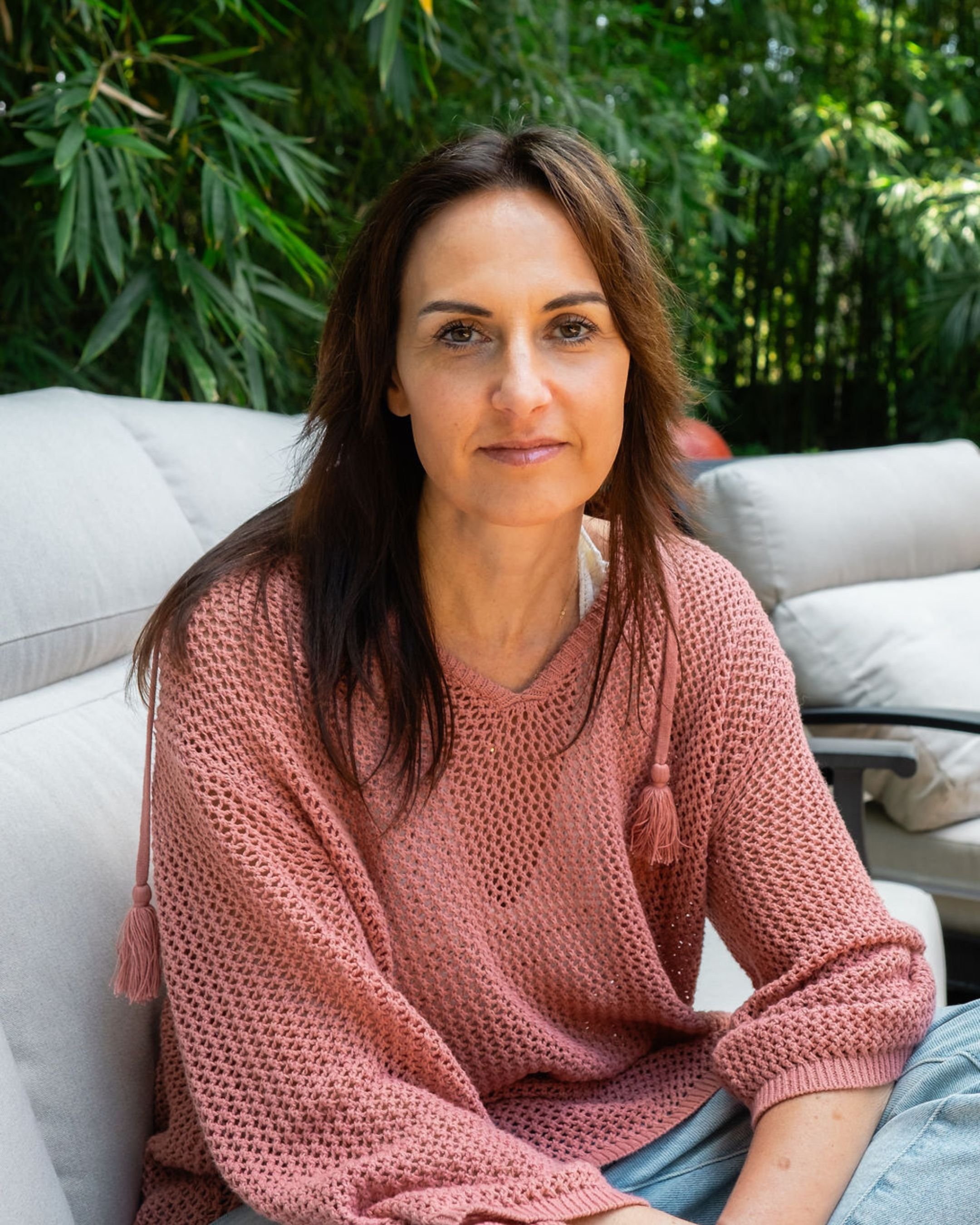 A woman with brown hair sitting outside on a white couch, wearing a pink knit sweater, with green foliage in the background.
