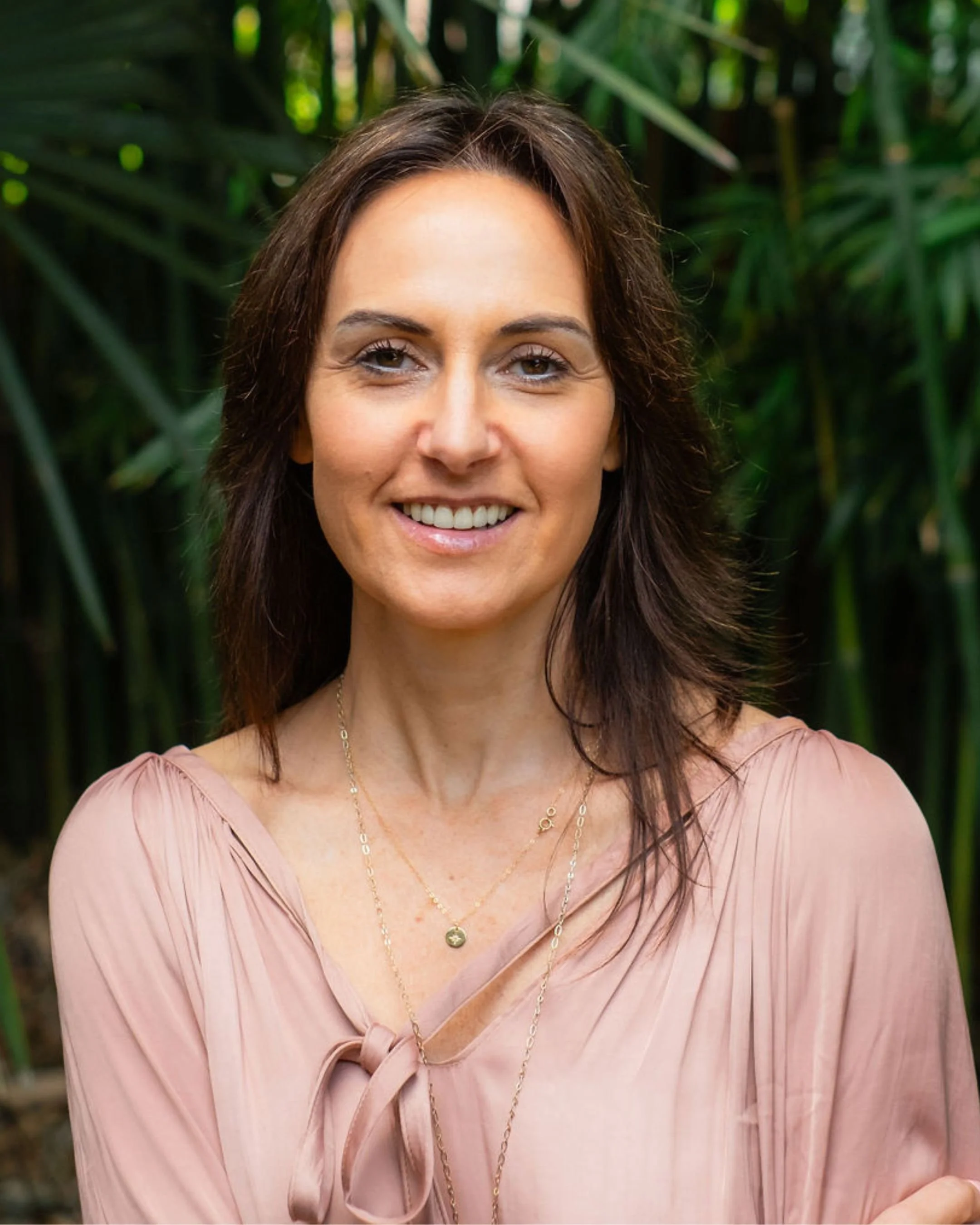 A woman with shoulder-length dark brown hair smiling outdoors with green foliage in the background.