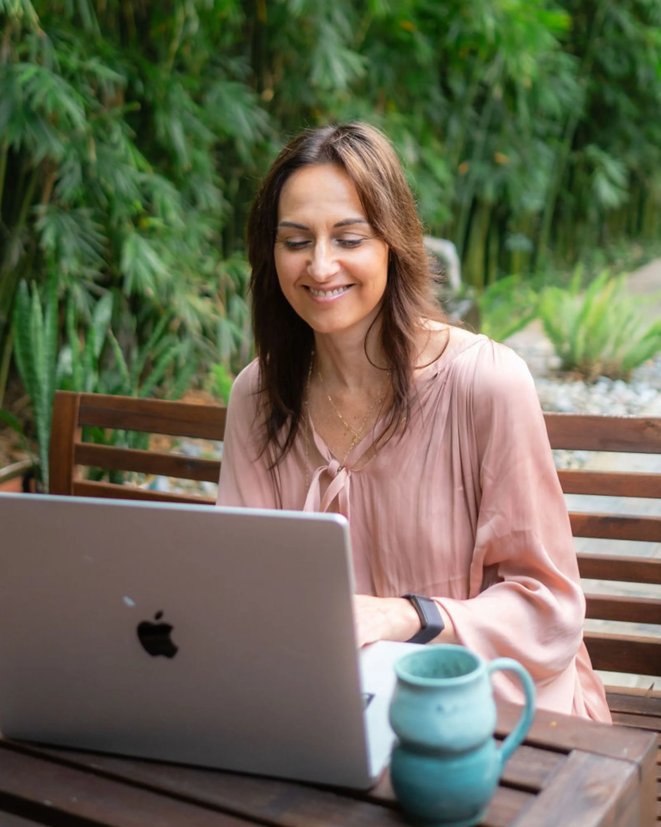 Woman sitting outdoors at a wooden table using a laptop, with greenery in the background, and a teal ceramic mug nearby.