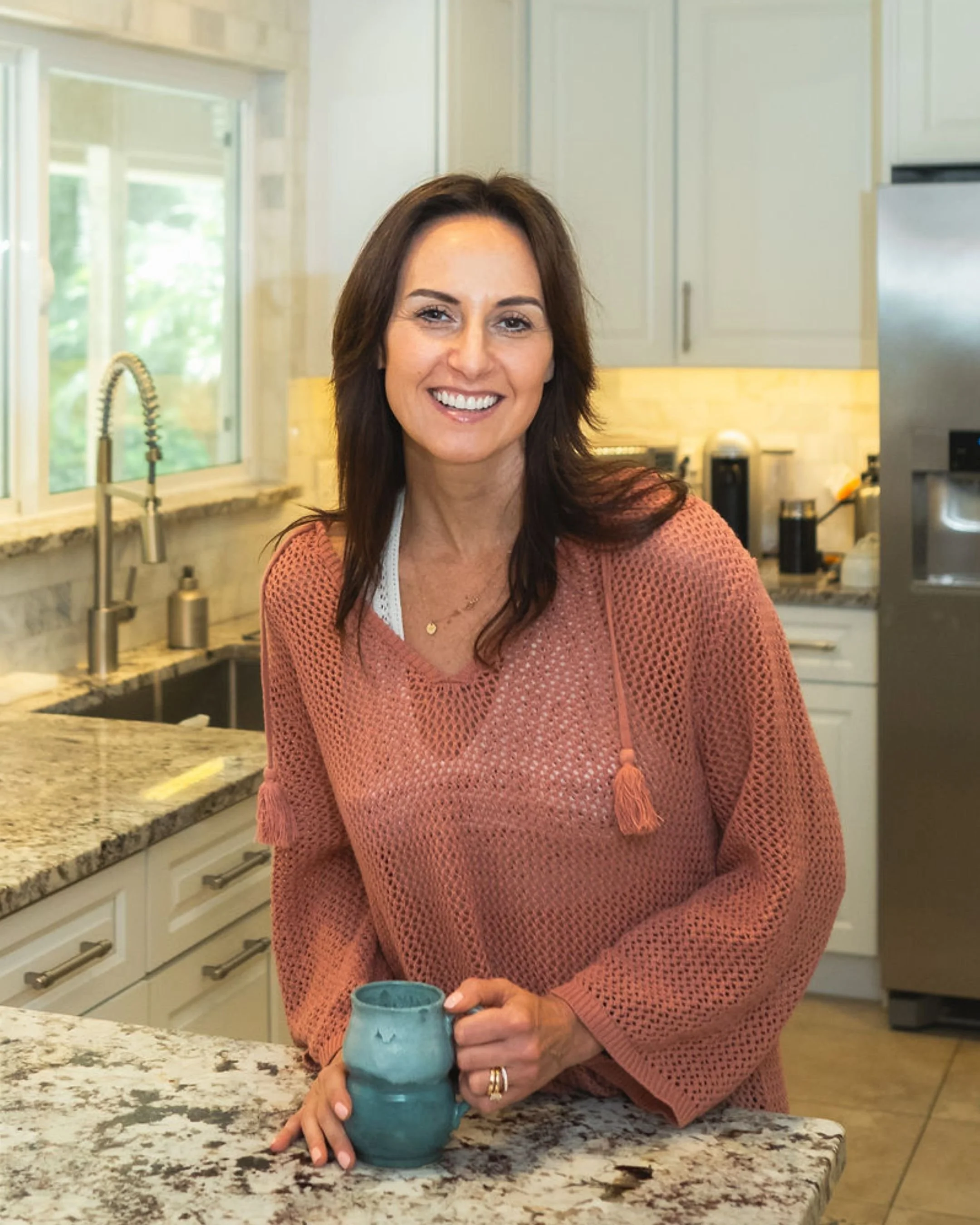 A woman smiling in a kitchen, holding a blue mug, wearing a pink knit sweater, with a window and kitchen appliances in the background.