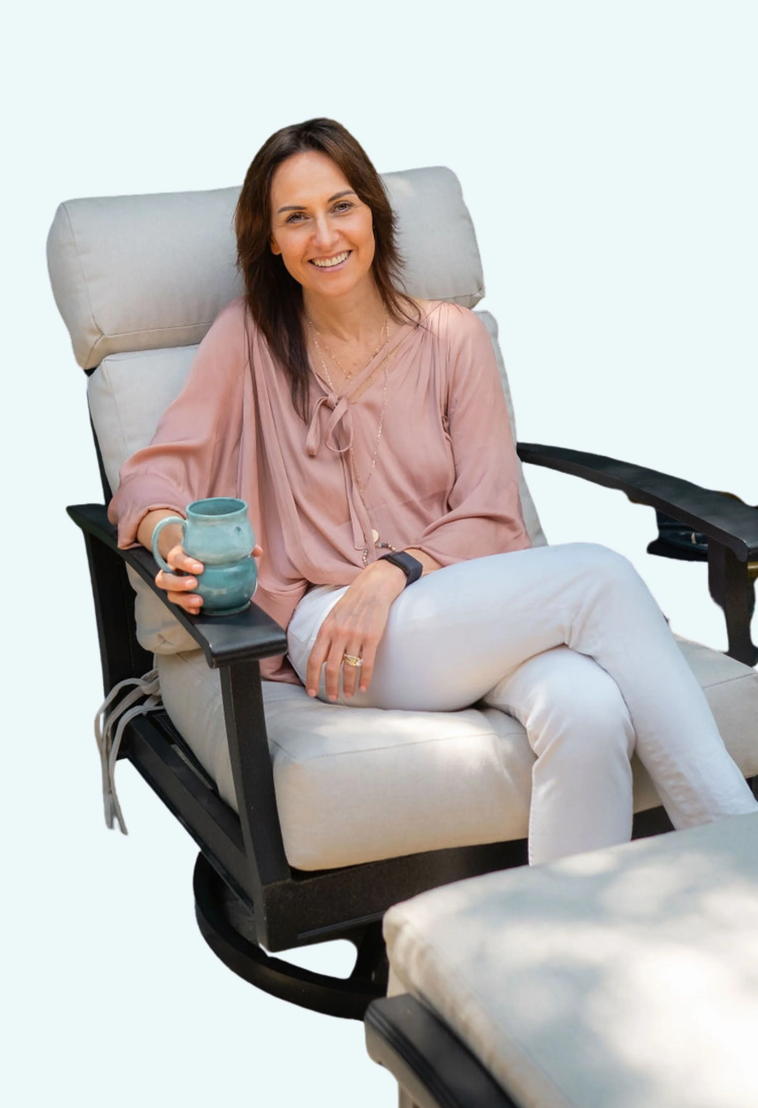 A woman sitting in a beige recliner, smiling and holding a blue ceramic mug, wearing a light pink blouse and white pants.