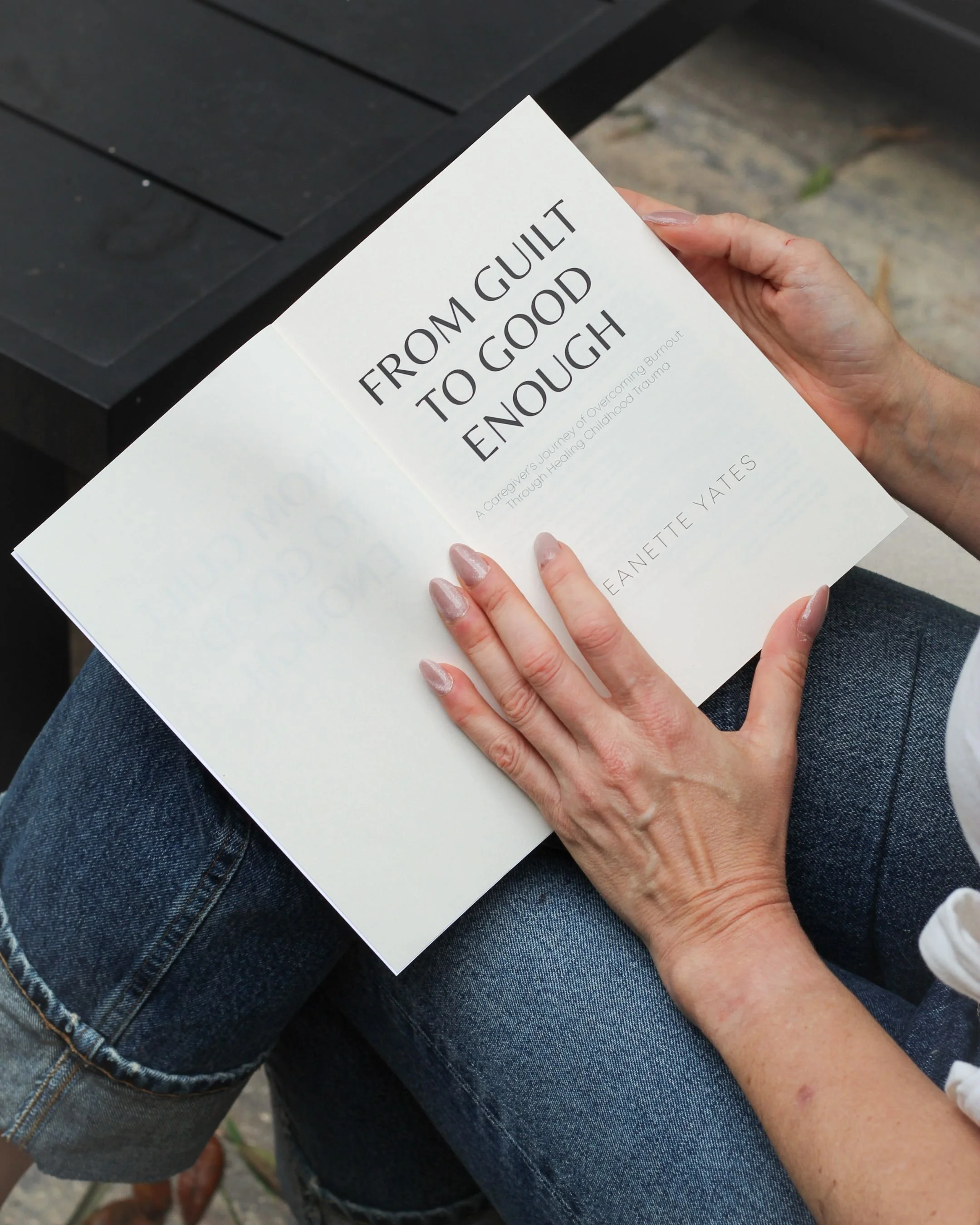 A person sitting outside is holding a book titled 'From Guilt to Good Enough' by Jeanette Yates. The person has painted nails and is wearing denim jeans.