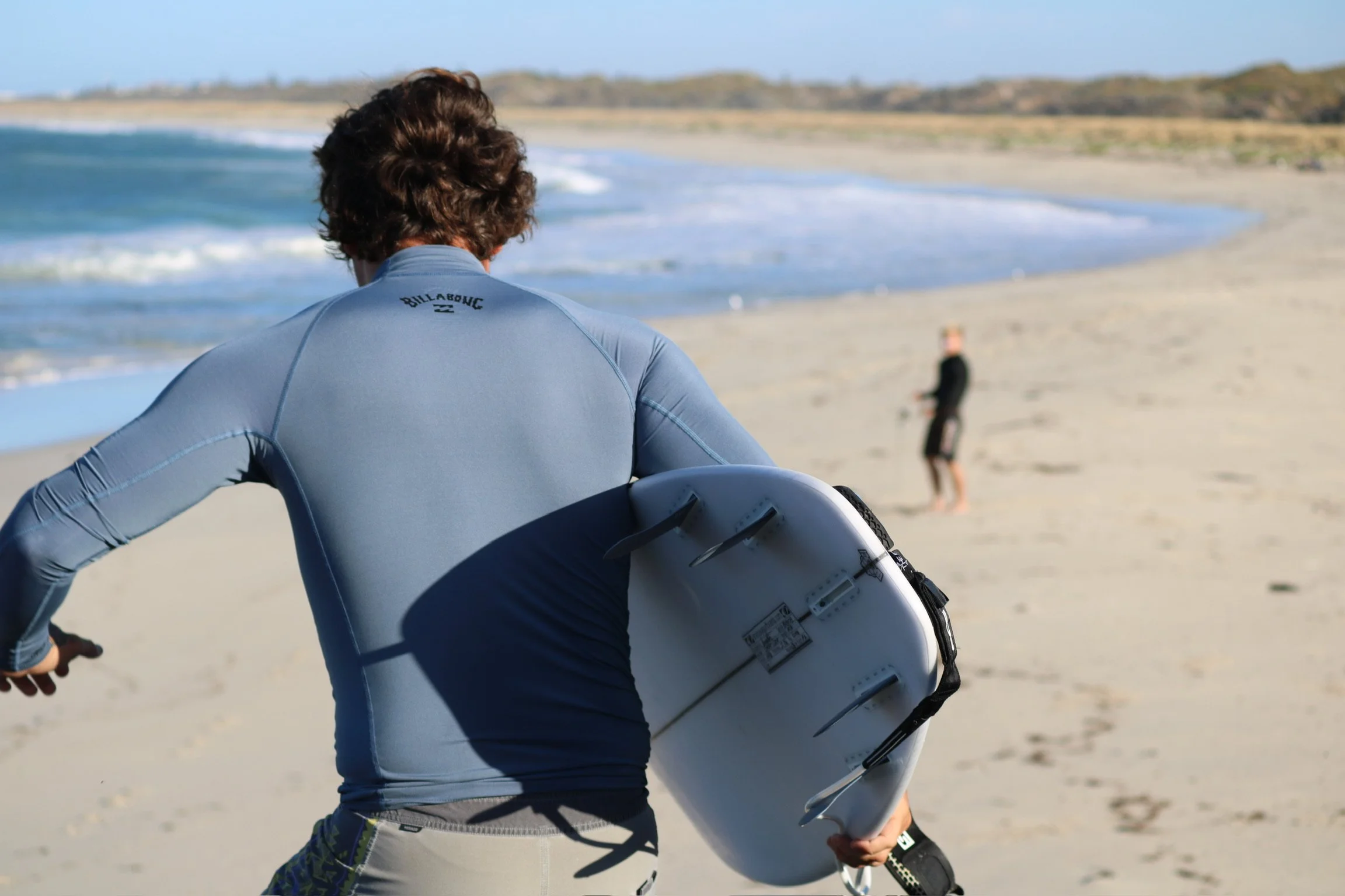 Person holding a surfboard walking on the beach towards the water with another person in the background.