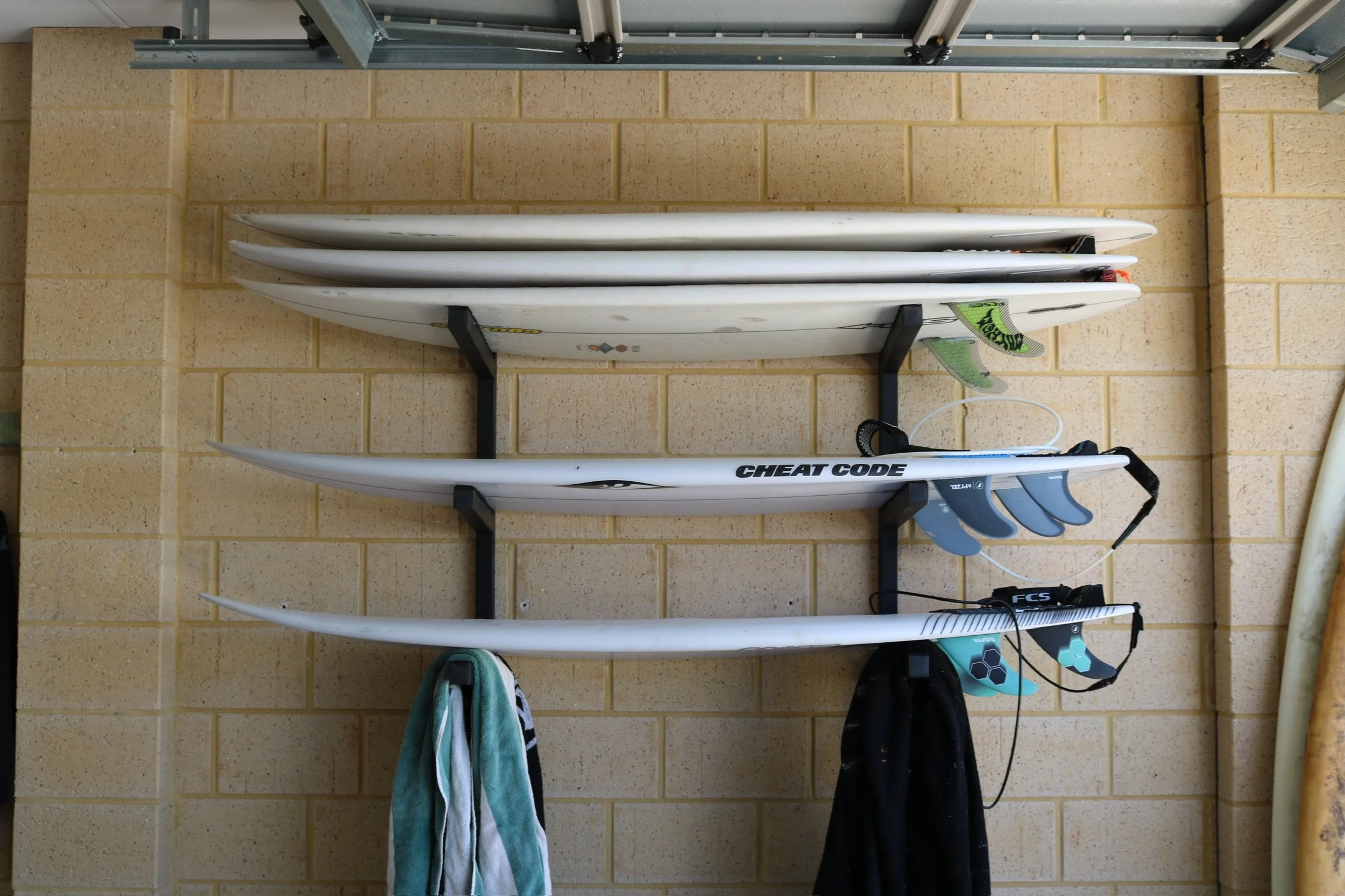 Four white surfboards with fins, mounted on a black rack on a brick wall in a garage. Two towels hang below the rack, and surfing accessories such as fins and a leash are visible.