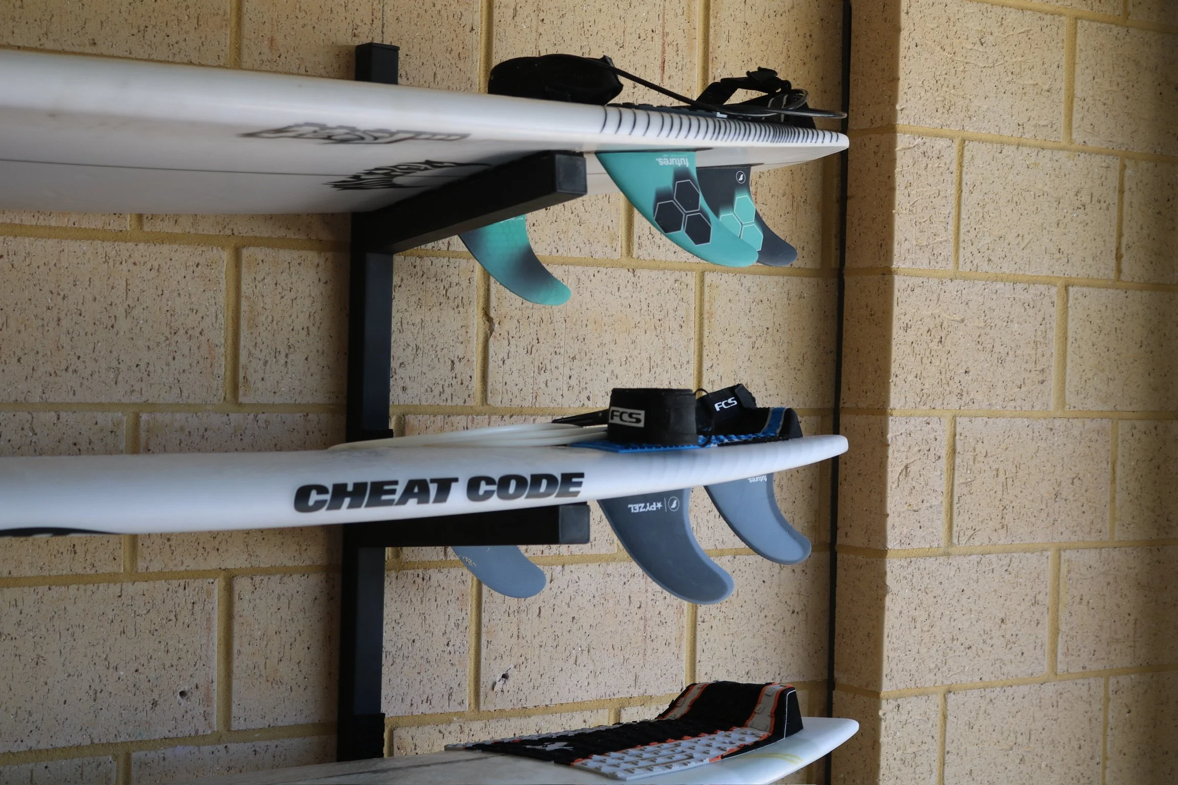 Four surfboards mounted on a wall rack against a yellow brick wall, with one surfboard featuring a