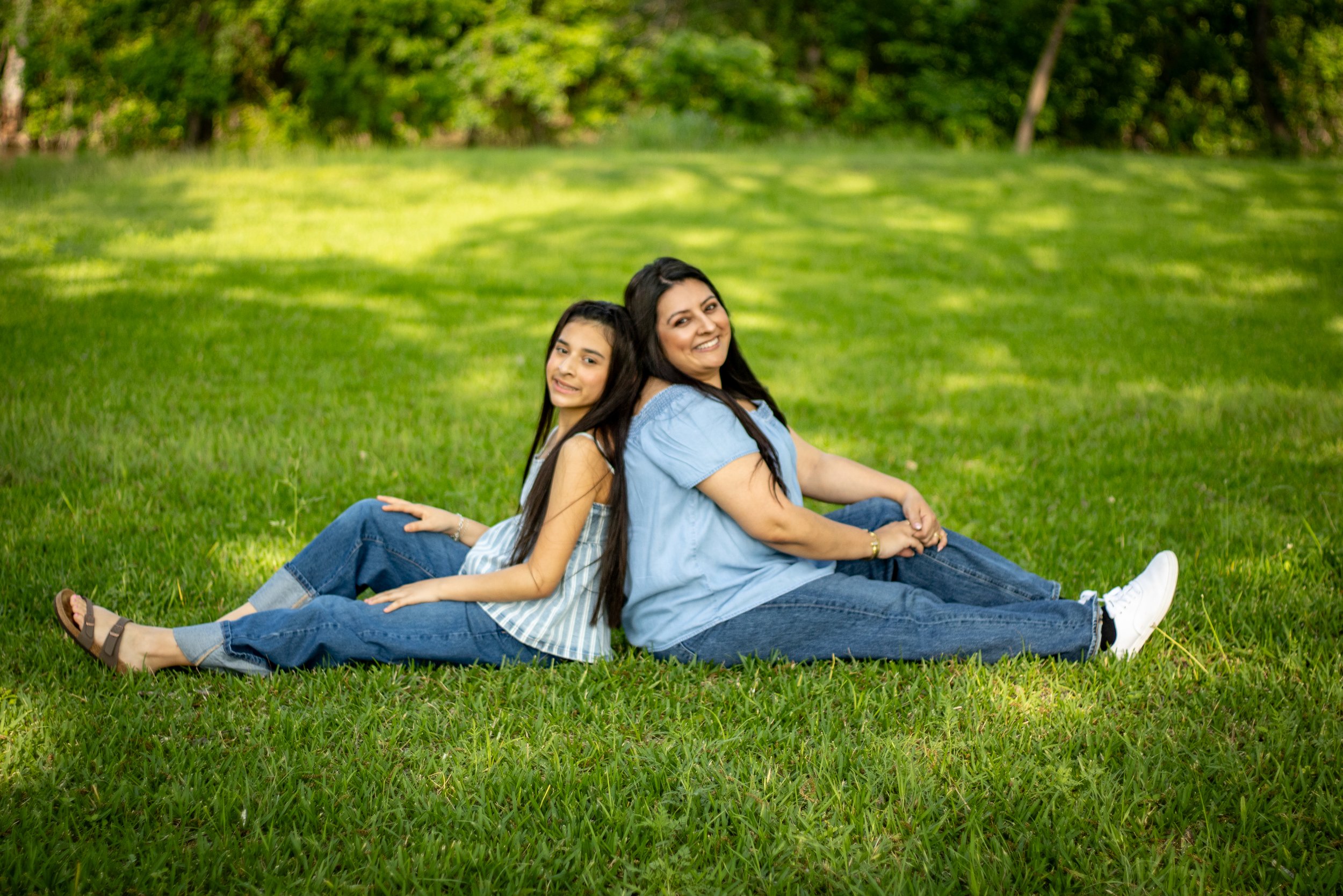 A woman and a girl sitting on grass in a park, back-to-back, smiling at the camera.