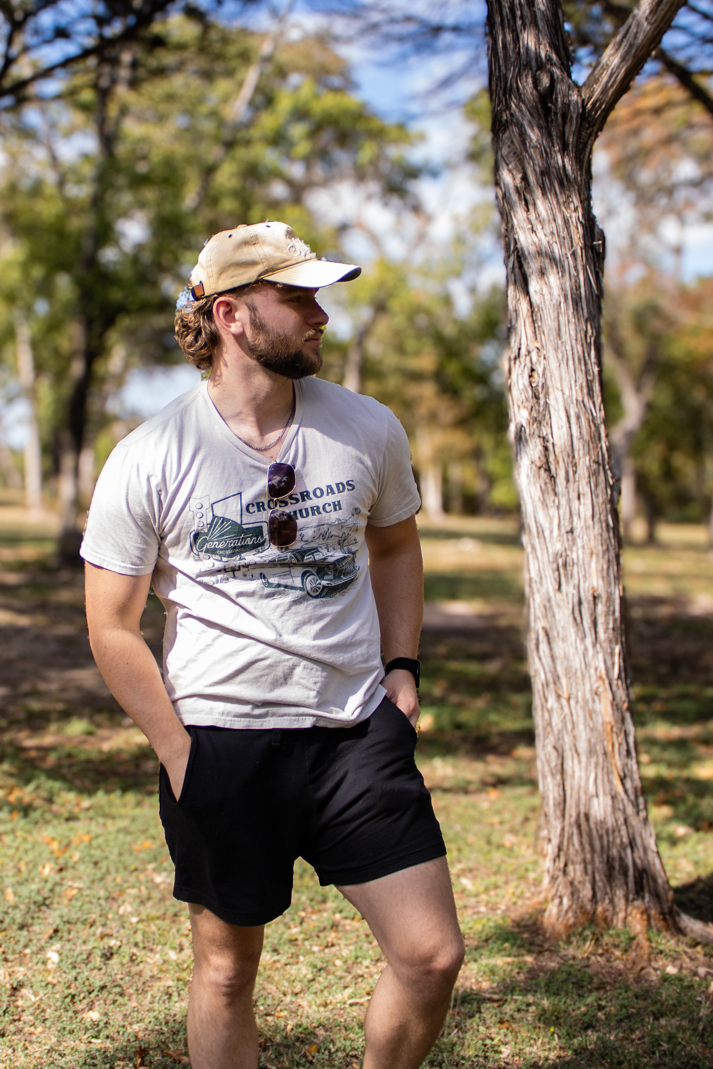 A young man with a beard, wearing a beige cap, white t-shirt, and black shorts, standing outdoors near a tree in a park during daytime, with other trees and a blue sky in the background.