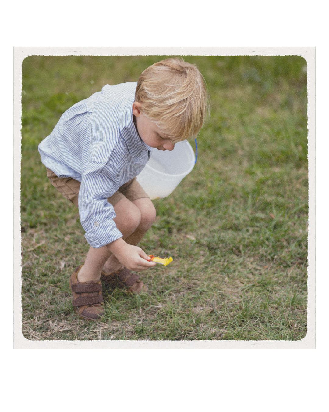 A young boy with blond hair wearing a striped shirt and shorts crouches on grass, looking at the ground while holding a small yellow toy.