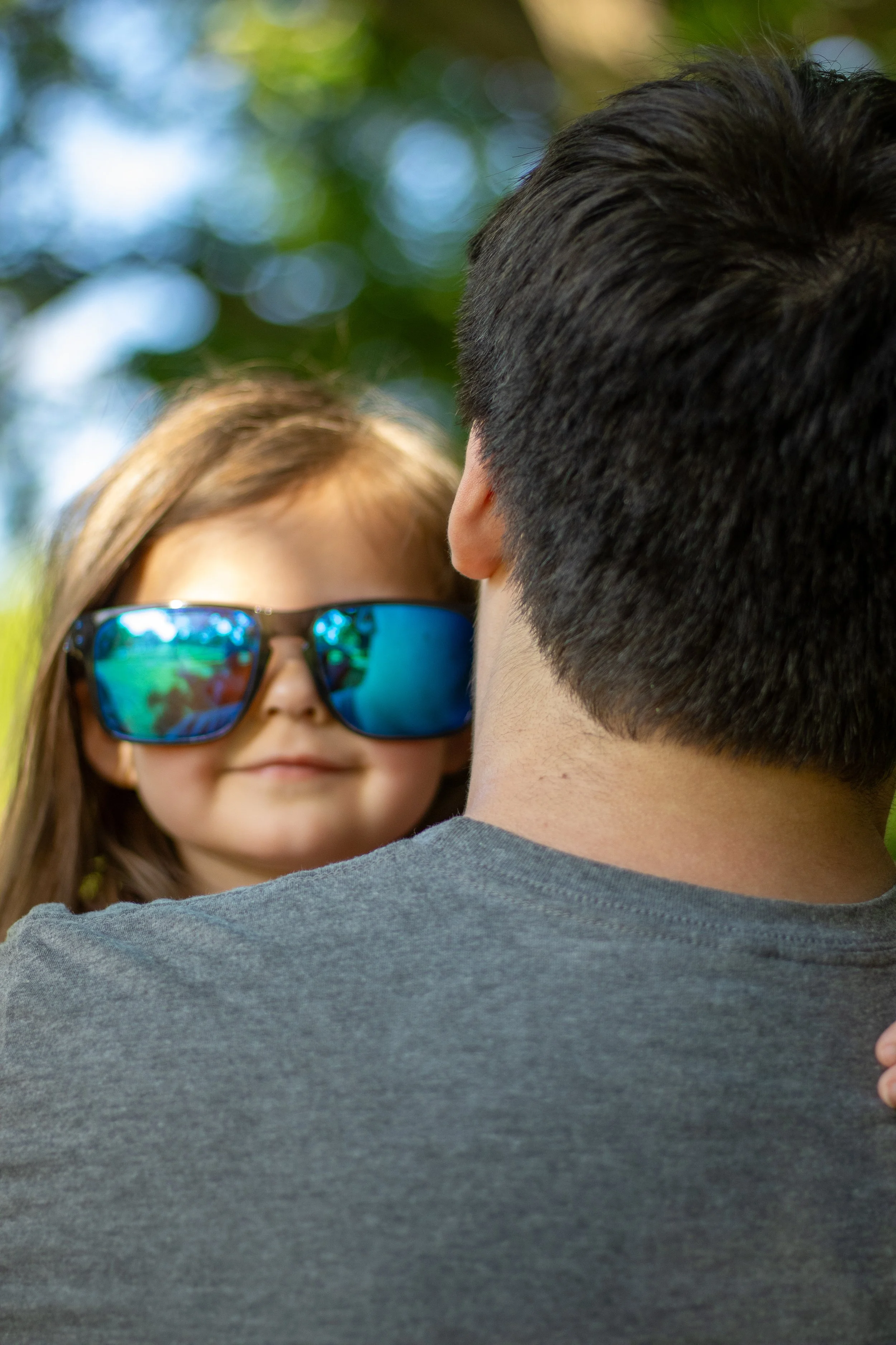 A woman with long blonde hair wearing sunglasses with reflective blue lenses stands behind a man with dark hair, outdoors with green leaves and sunlight in the background.