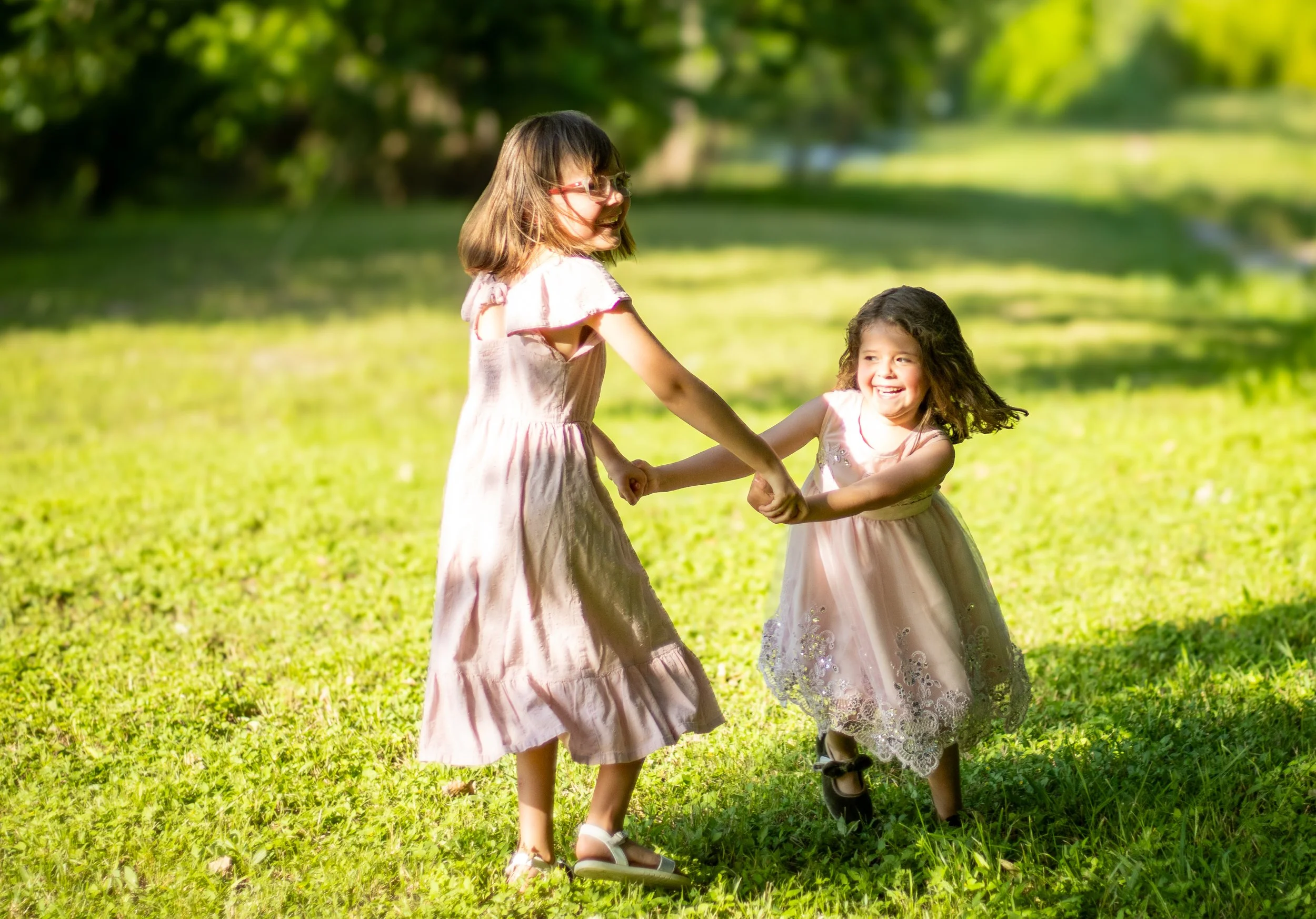 Two young girls holding hands and spinning around outdoors in a grassy park on a sunny day, smiling and enjoying themselves.