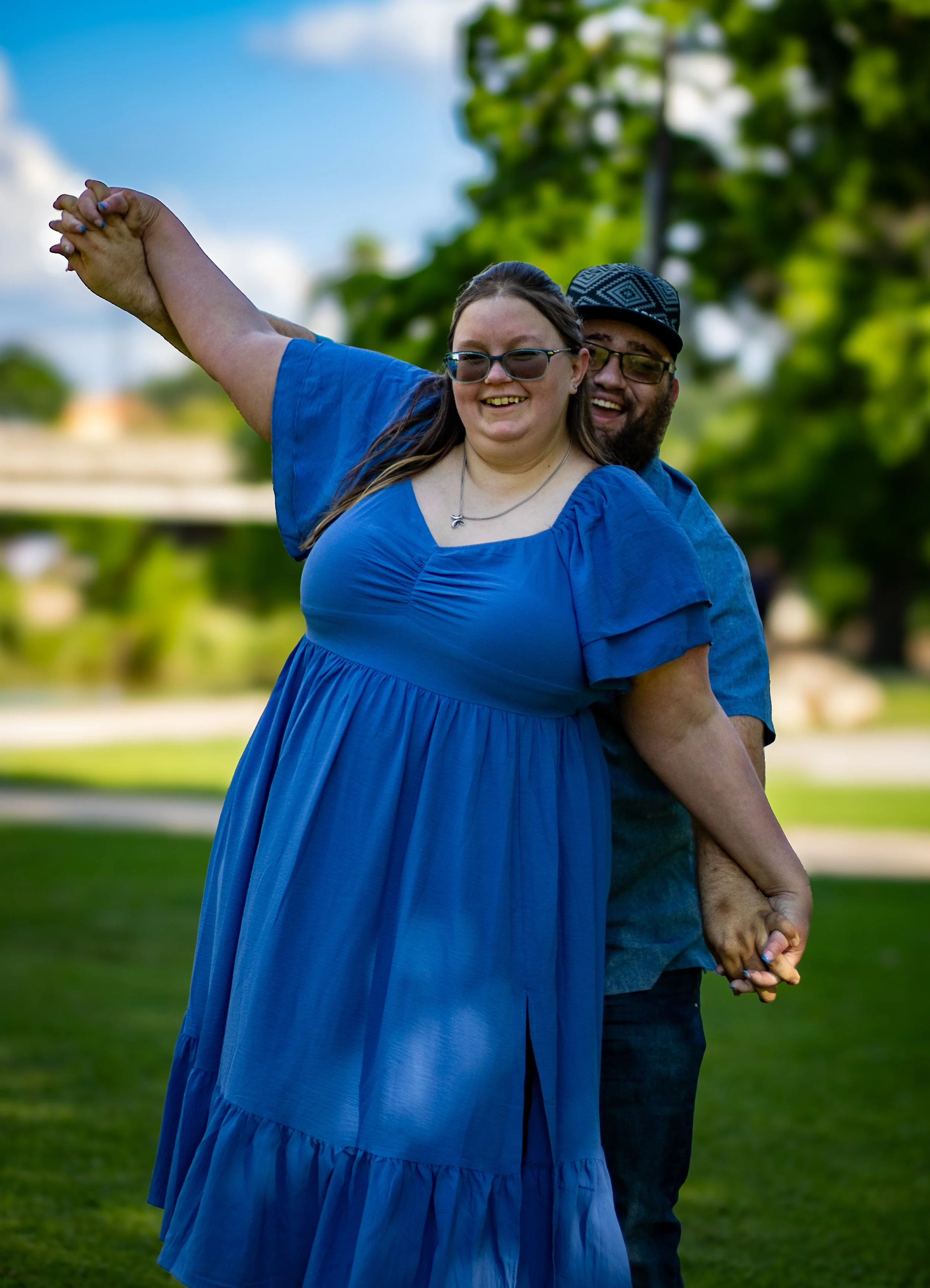A smiling woman in a blue dress and sunglasses is holding hands with a smiling man behind her, both standing in a park with green trees and a blue sky.