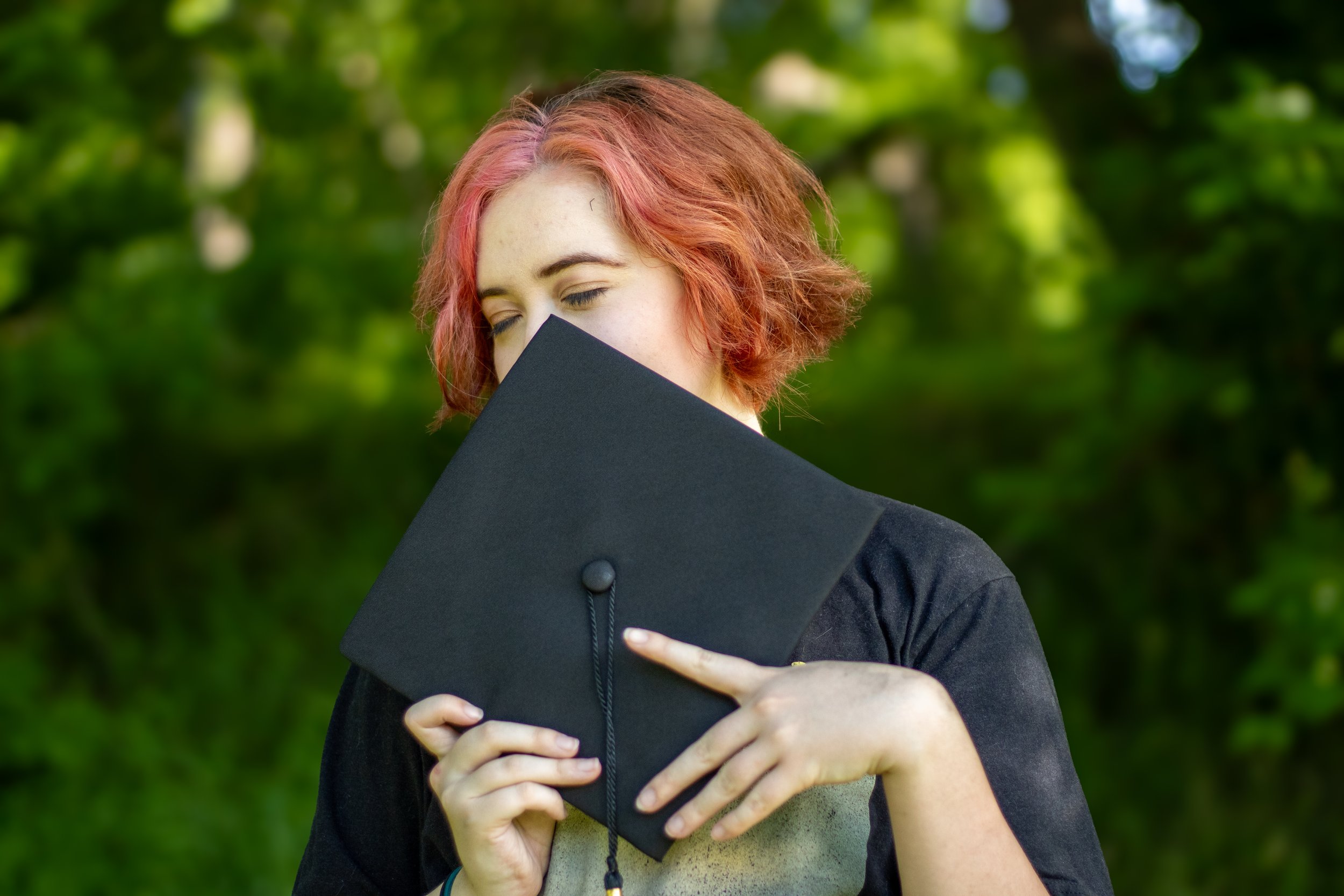 A young woman with red hair holding a diploma, eyes closed, outdoors with green foliage in the background.