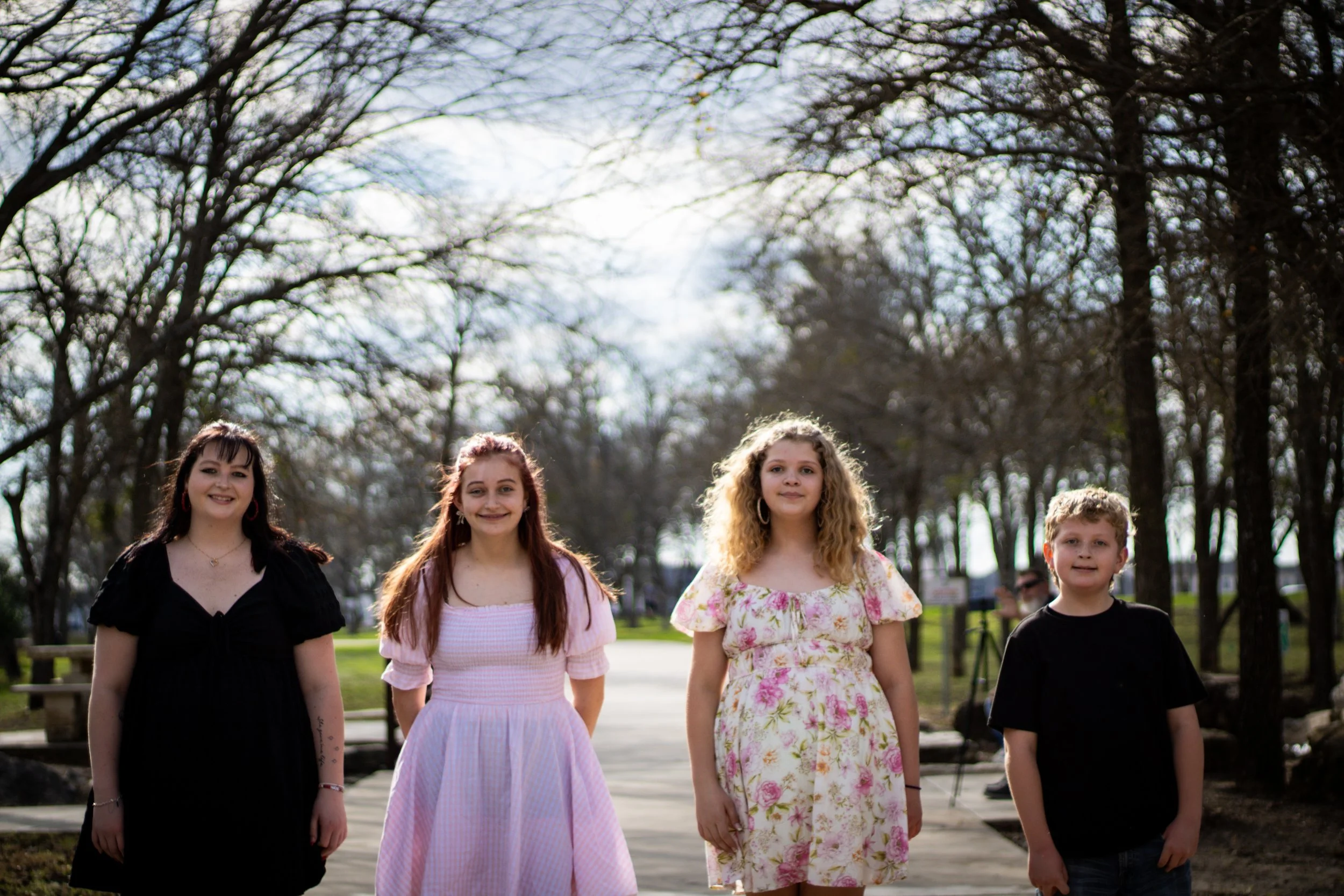 Five young people standing outdoors in a park with trees, smiling at the camera.