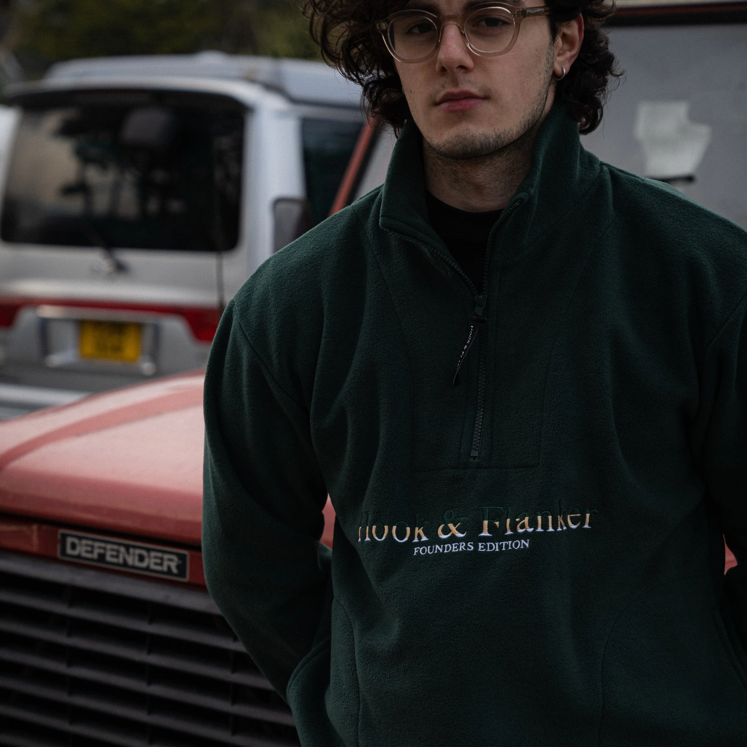 A young man with curly brown hair and glasses, wearing a dark green jacket with embroidered text 'Hook & Flanker Founder’s Edition', standing in front of a red vehicle with a license plate, and a white vehicle with a yellow license plate in the background.