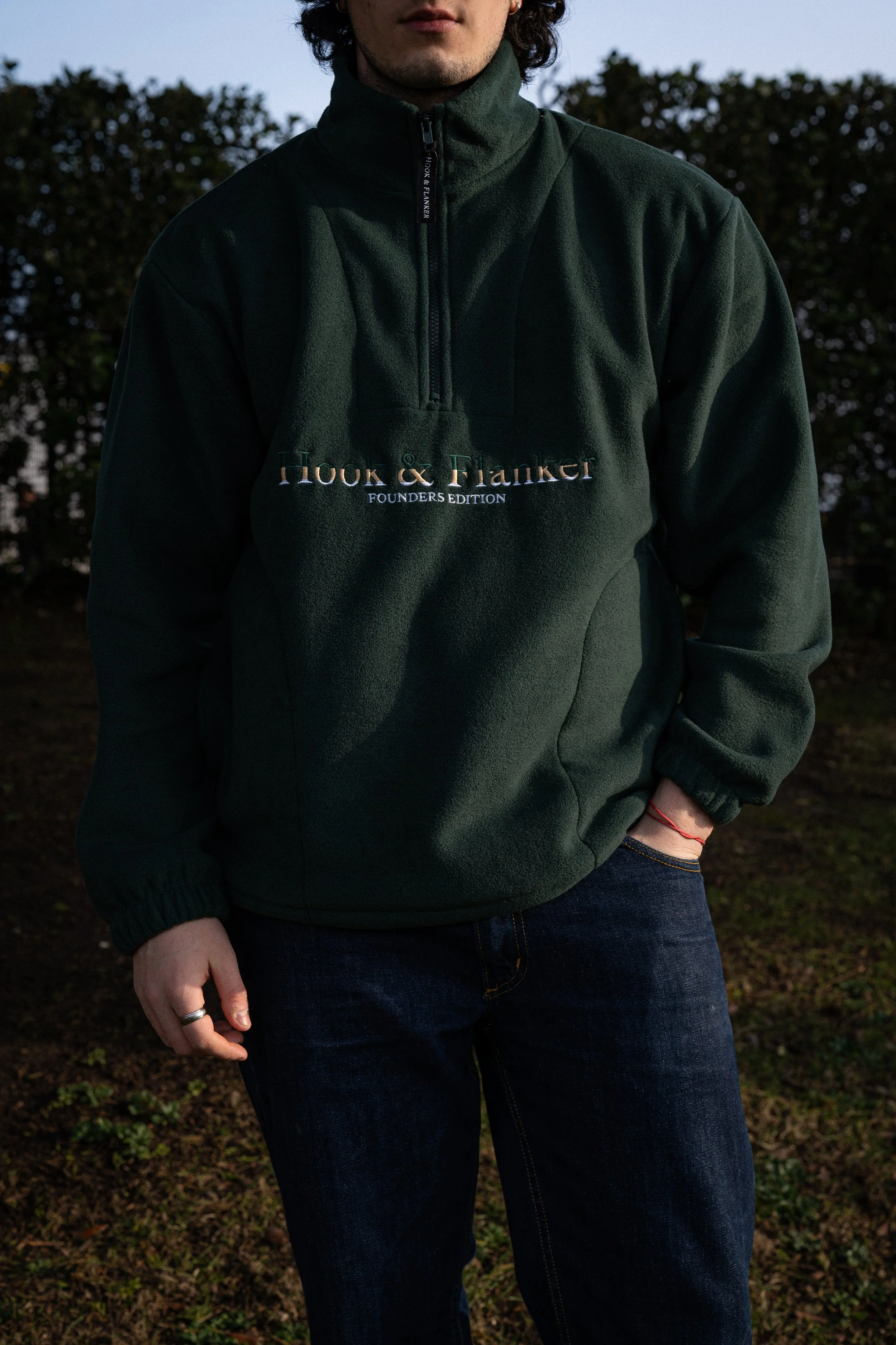Person wearing a dark green Hook & Flanker fleece jacket with their hand in their pocket, standing outdoors during daylight.