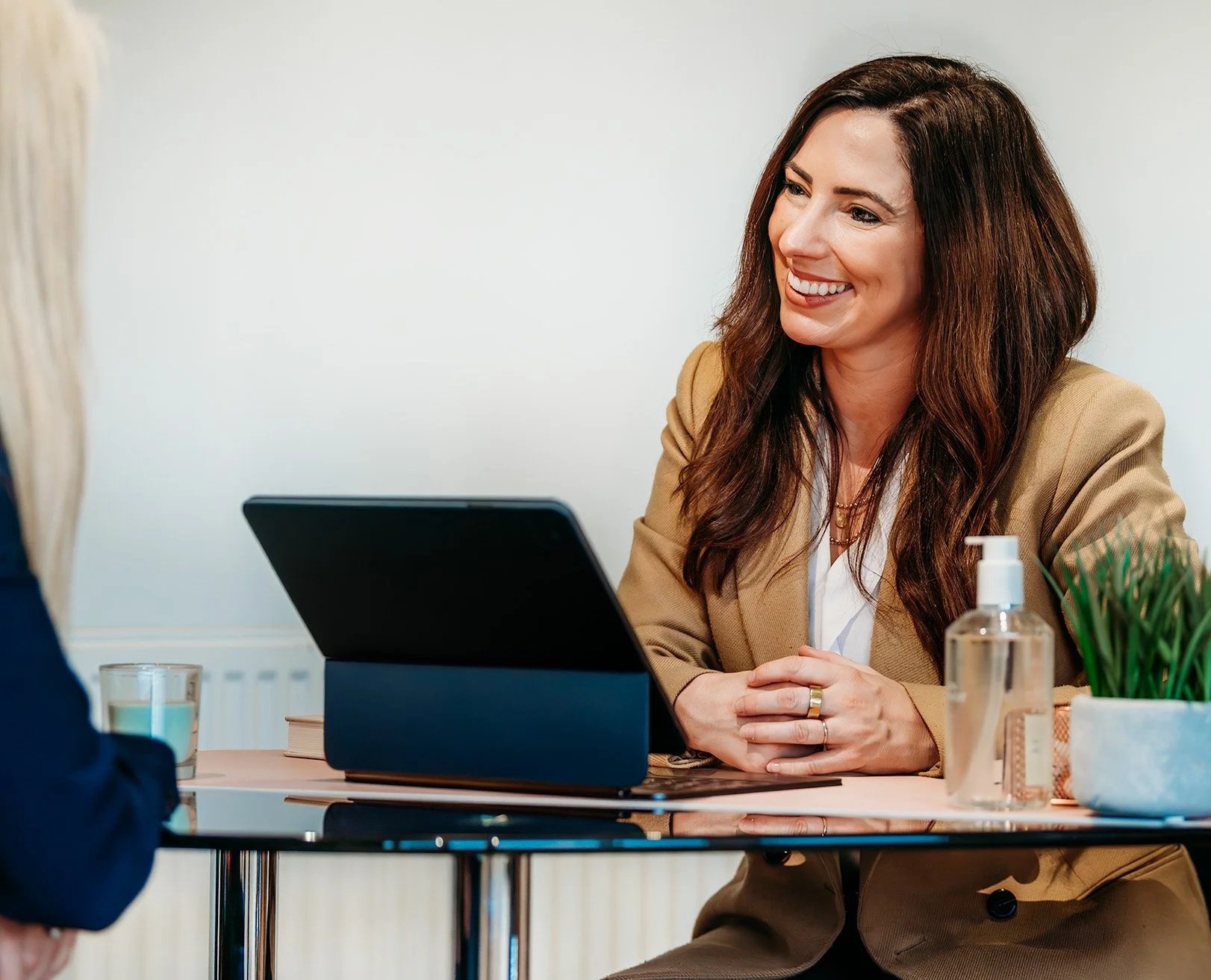A woman in a tan blazer smiling and talking with a client at a desk, with a laptop, hand sanitiser, a plant, and a glass of water on the table.