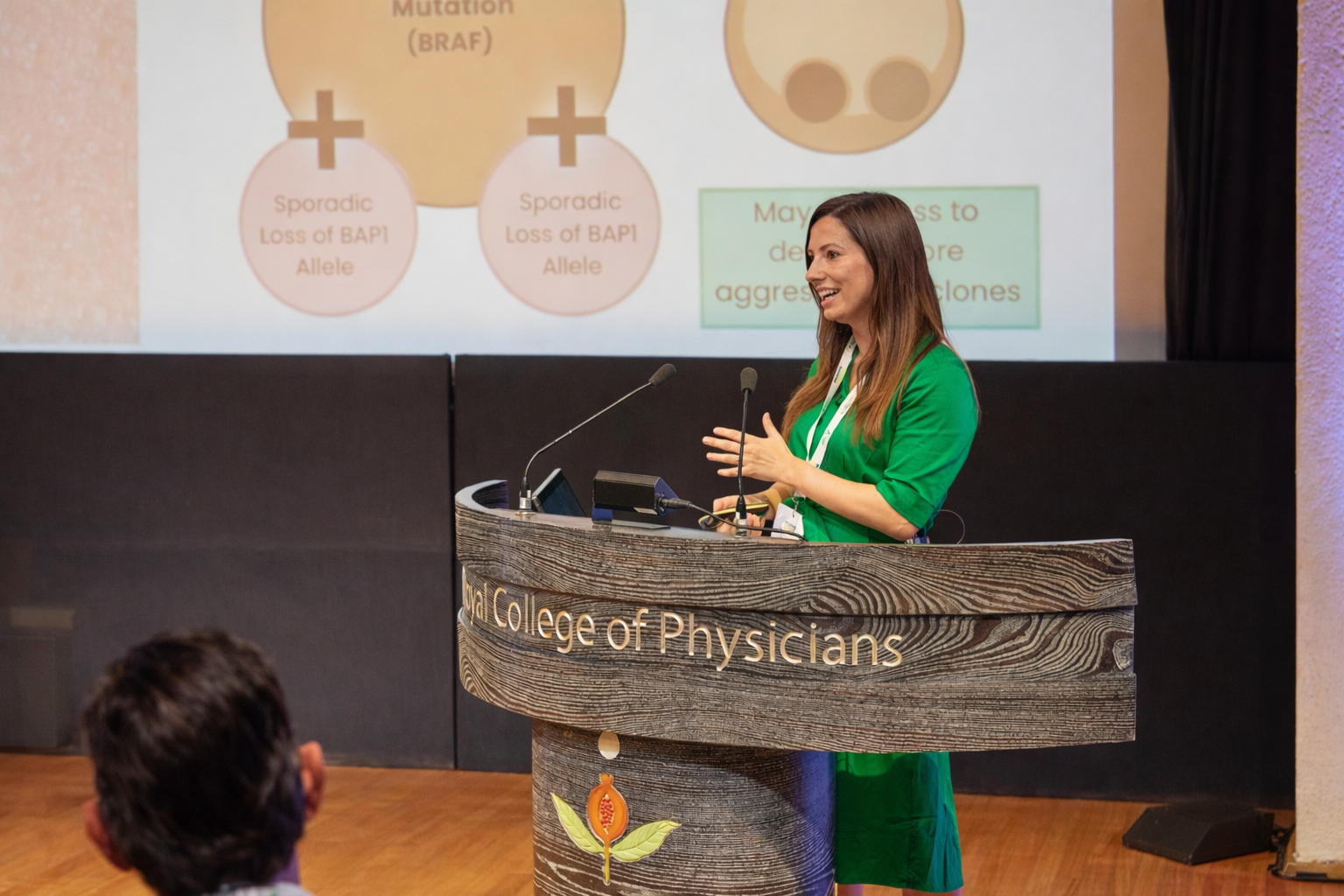 A woman in a green dress giving a presentation at the Central College of Physicians, standing behind a wooden podium with microphones, with a slide projected behind her showing diagram related to genetics.