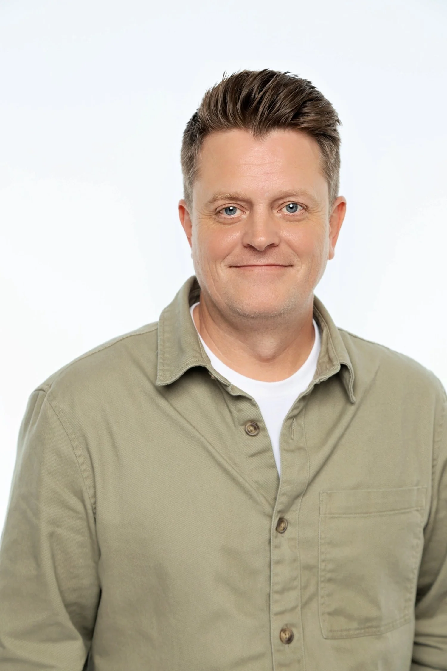 A man with short, styled brown hair, blue eyes, and a light complexion, wearing a beige button-up shirt over a white undershirt, standing against a plain white background.
