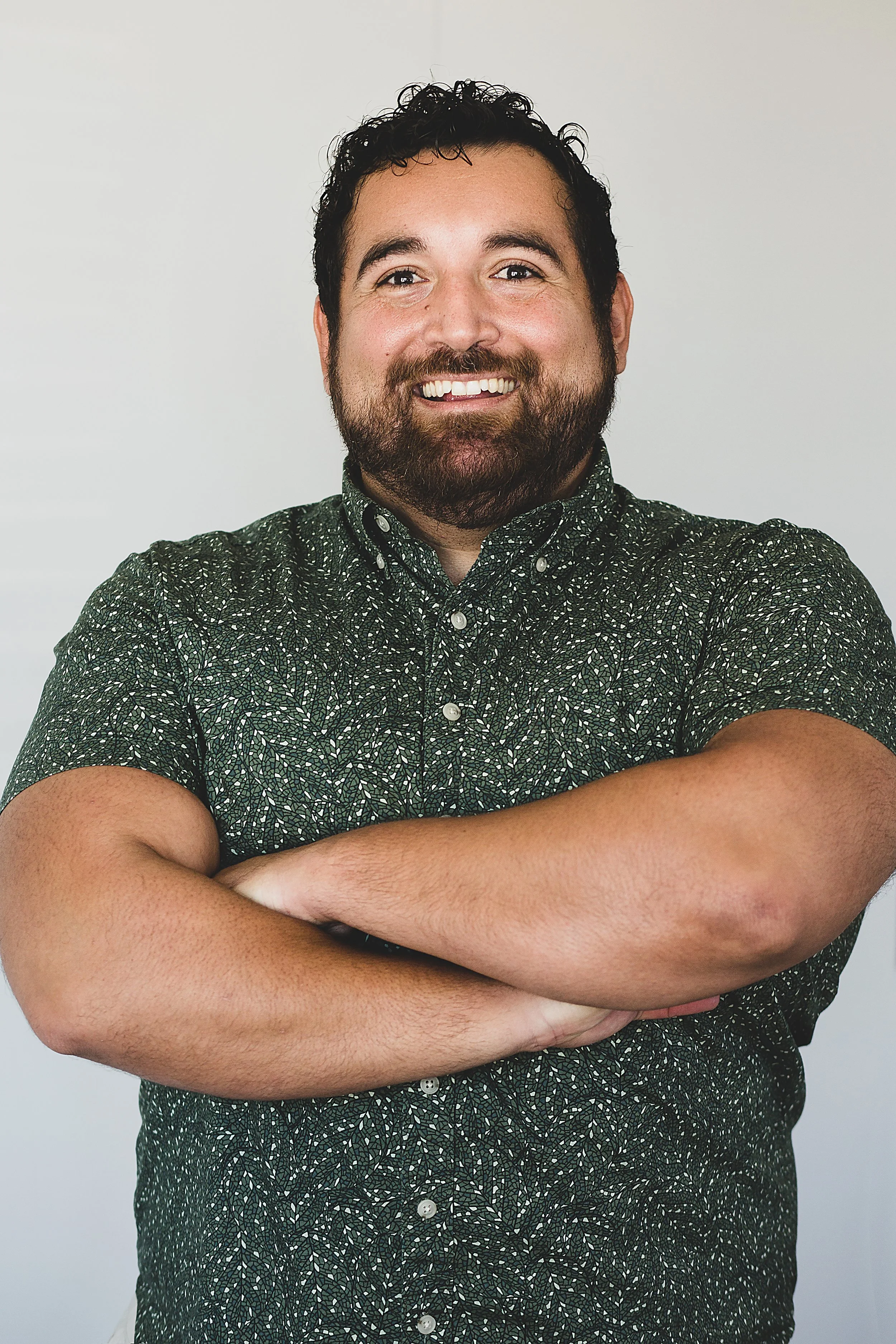A smiling man with dark curly hair and a beard, wearing a green patterned button-up shirt, crossing his arms, standing against a plain light-colored background.