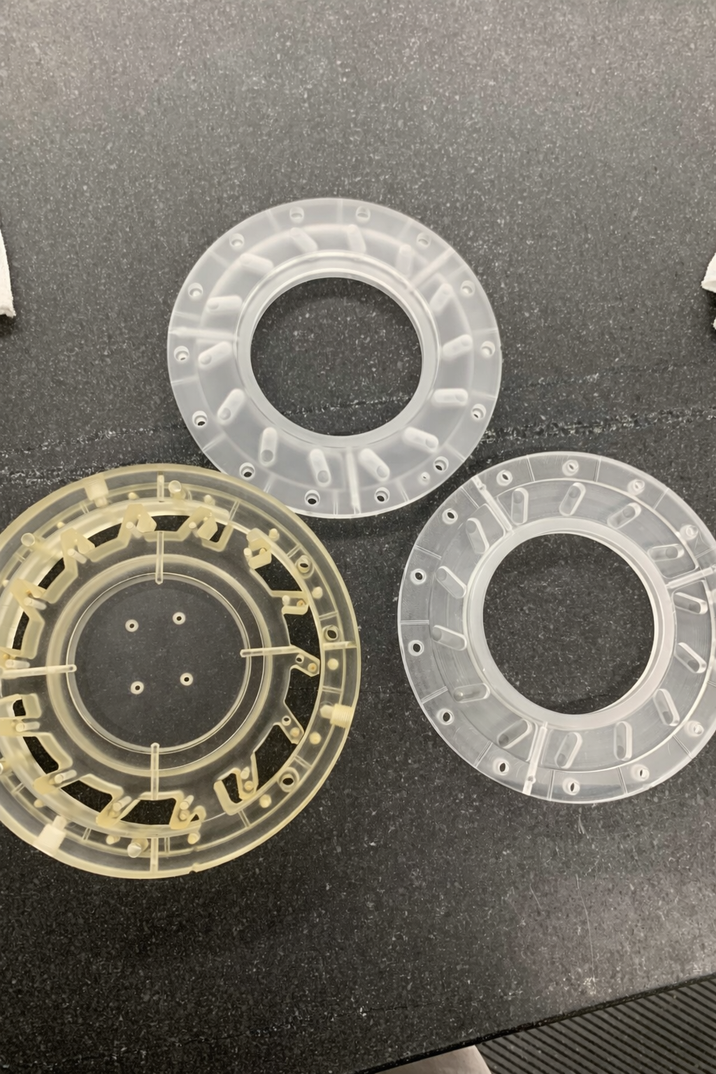 Three circular plastic components, two clear and one beige, placed on a black surface with specks.