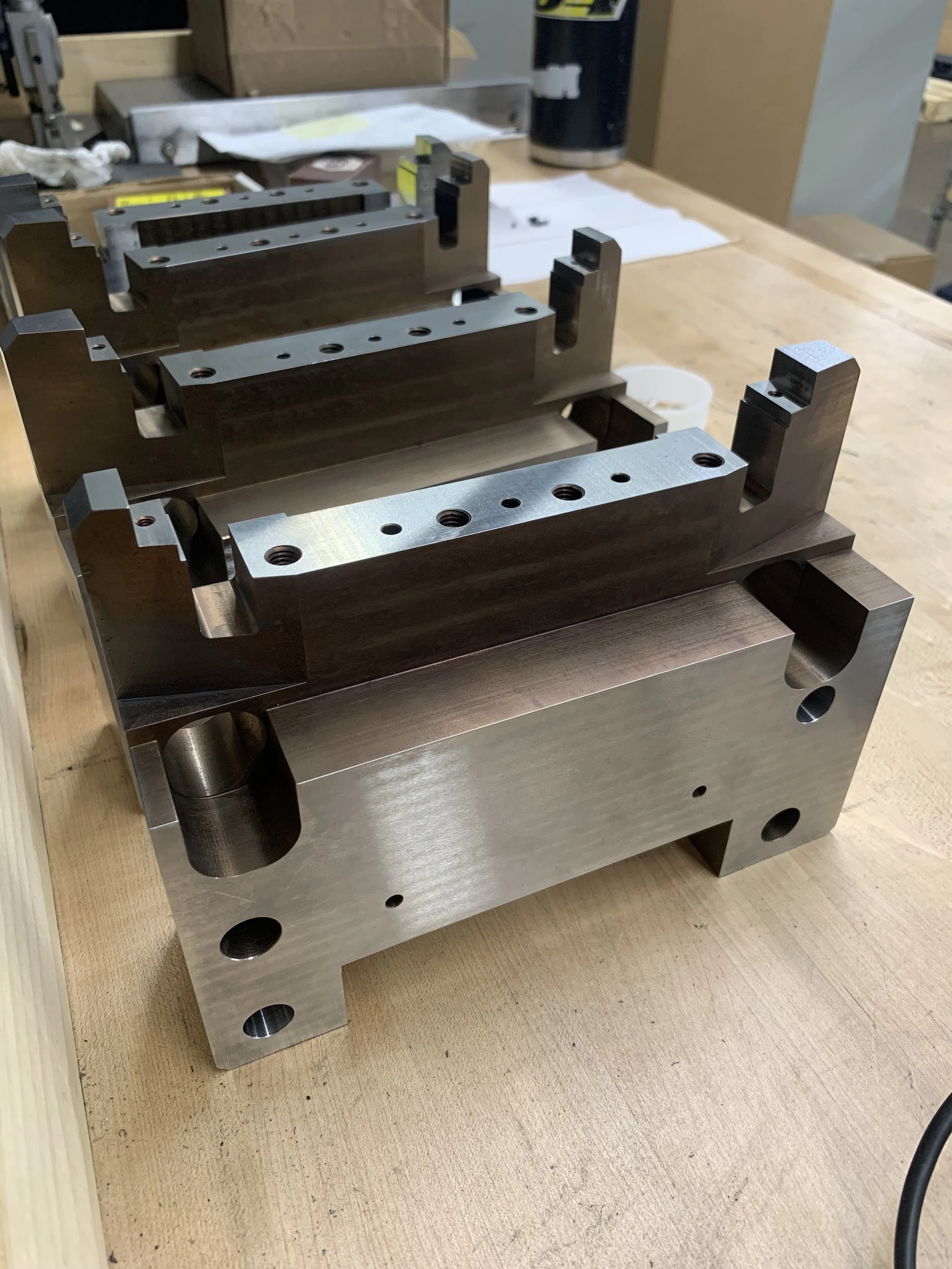 Three metallic machined parts, possibly mold or machine components, placed on a wooden workbench in an industrial or workshop setting.