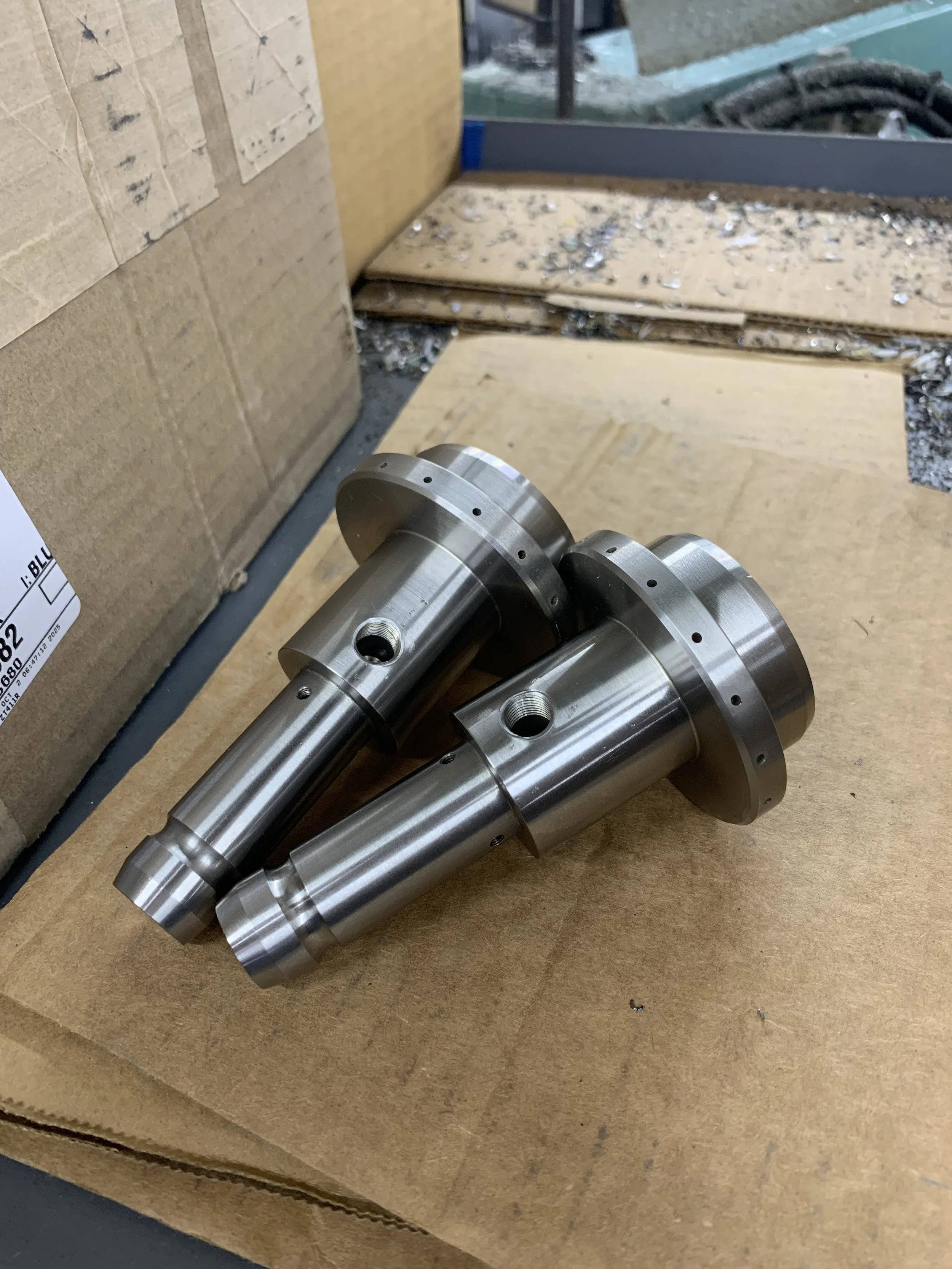 Two metal machined parts with cylindrical shapes and flanged ends placed on a cardboard surface, with a box and some debris in the background.